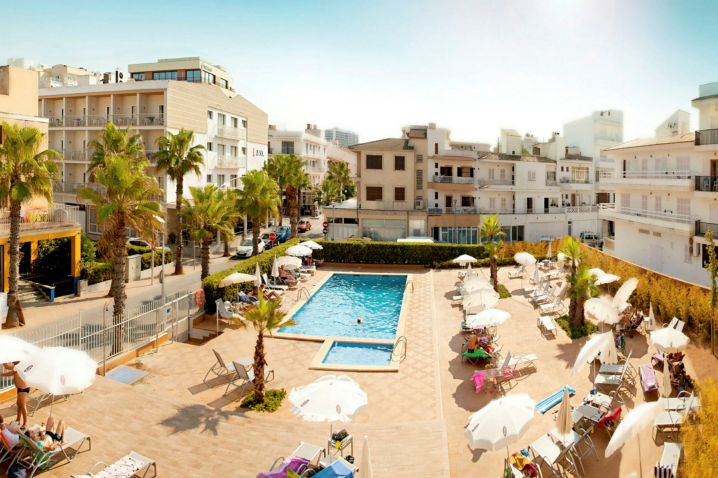 Hotellets poolområde med liggestole og parasoller omgivet af bygninger. (AI-genereret tekst)