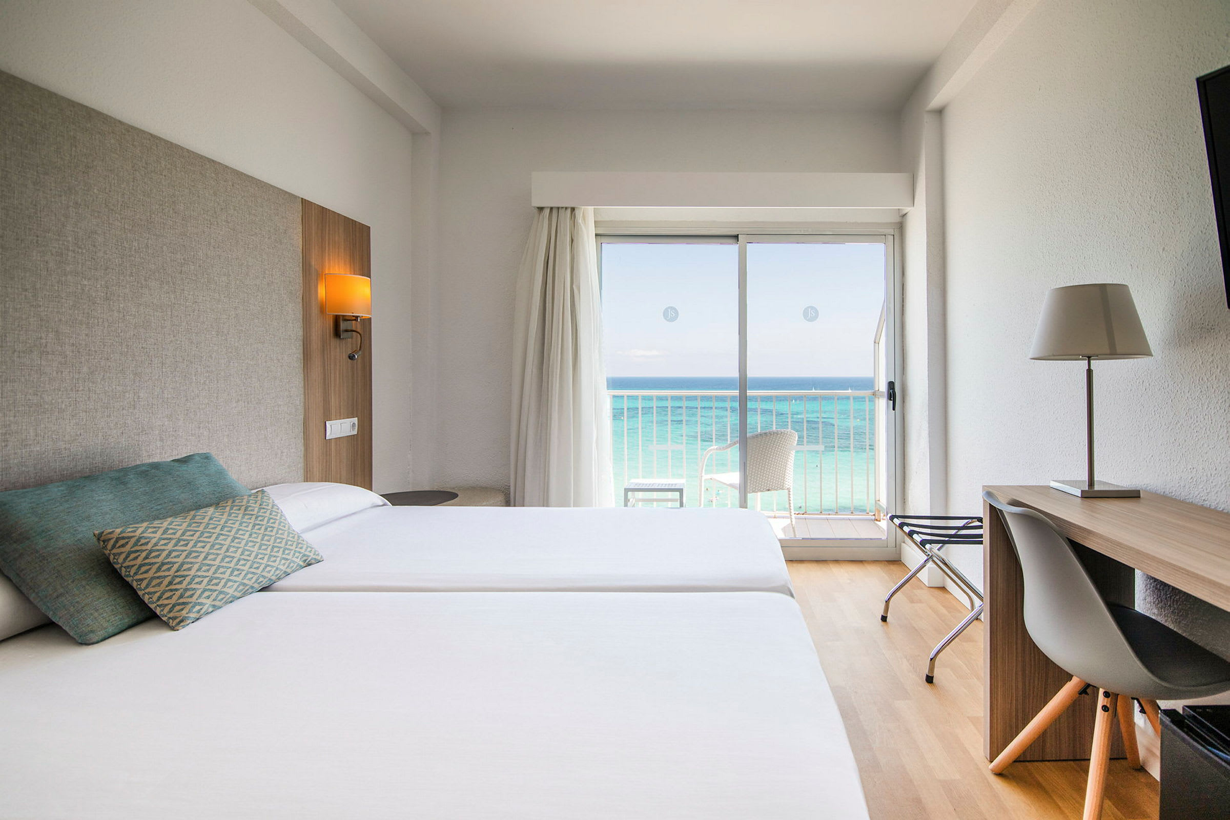 Hotelværelse med to senge, skrivebord og udsigt over havet gennem en glasdør. (AI-genereret tekst)