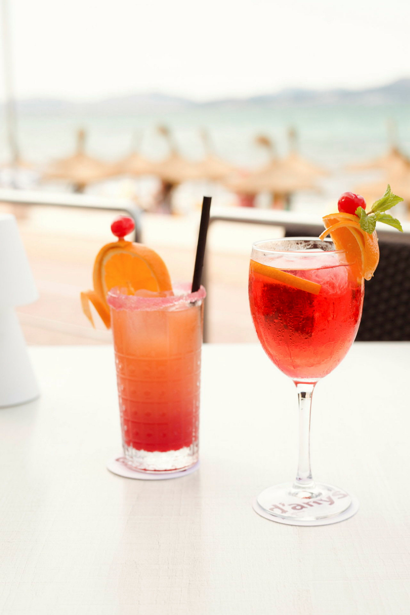 To farverige drinks dekoreret med frugt på et bord, med en sløret strand i baggrunden. (AI-genereret tekst)