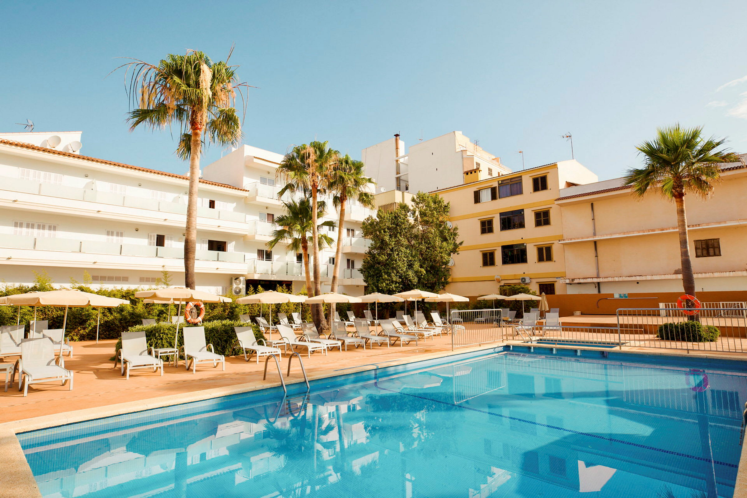 Udendørs billede af hotel med pool og solsenge, omgivet af bygninger og palmer. (AI-genereret tekst)