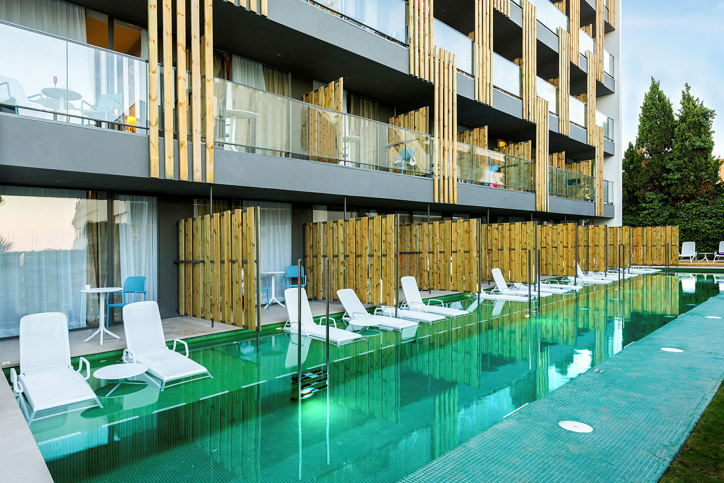 Billede af et hotel med direkte adgang til poolen fra visse værelser. Liggestole står ved poolkanten. (AI-genereret tekst)