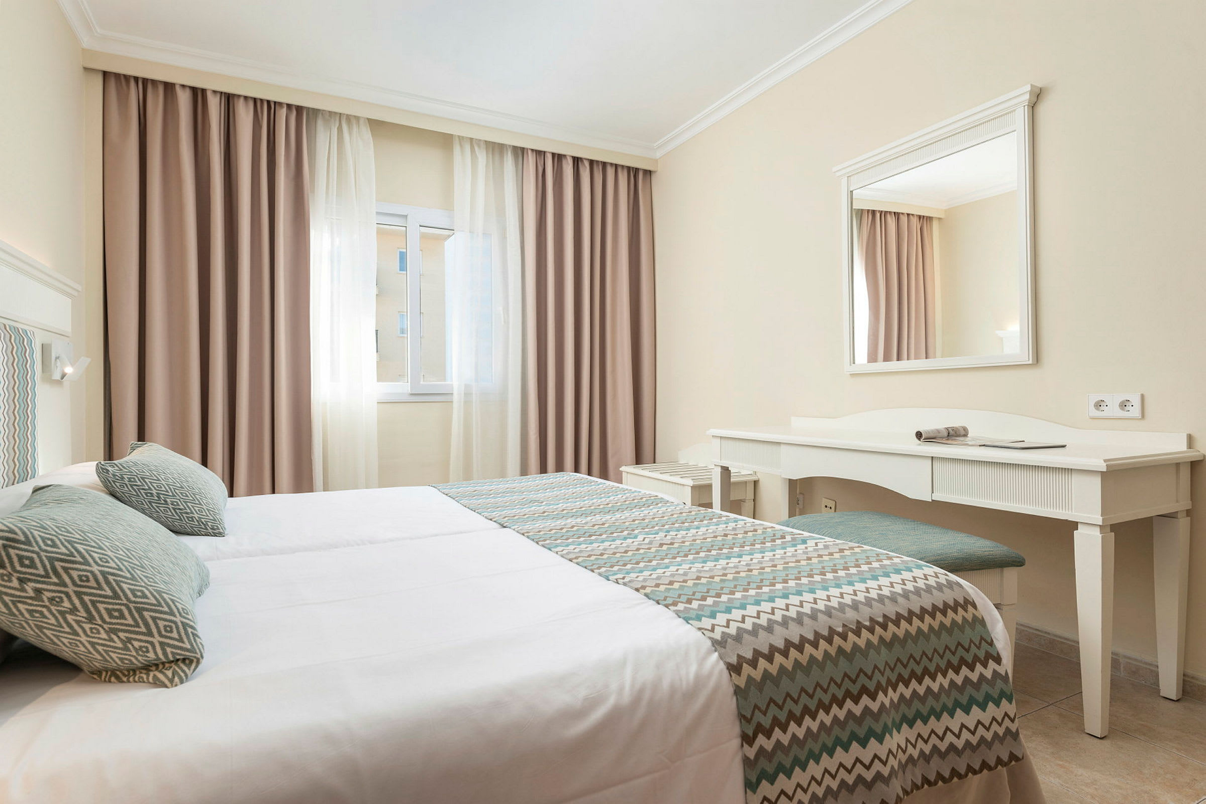 Hotelværelse med enkeltsenge, vindue med lyse gardiner og et skrivebord med spejl. (AI-genereret tekst)