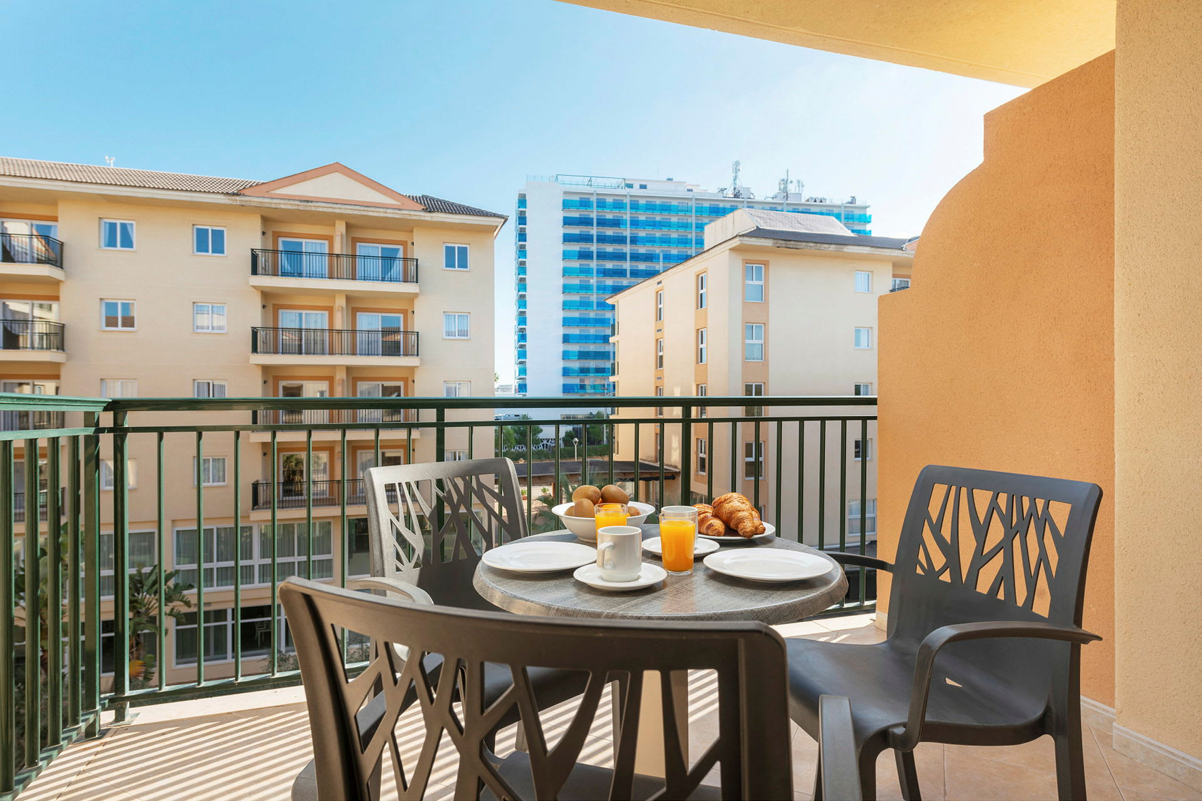 Udsigt fra balkon med møbler, morgenmad og omkringliggende bygninger. (AI-genereret tekst)