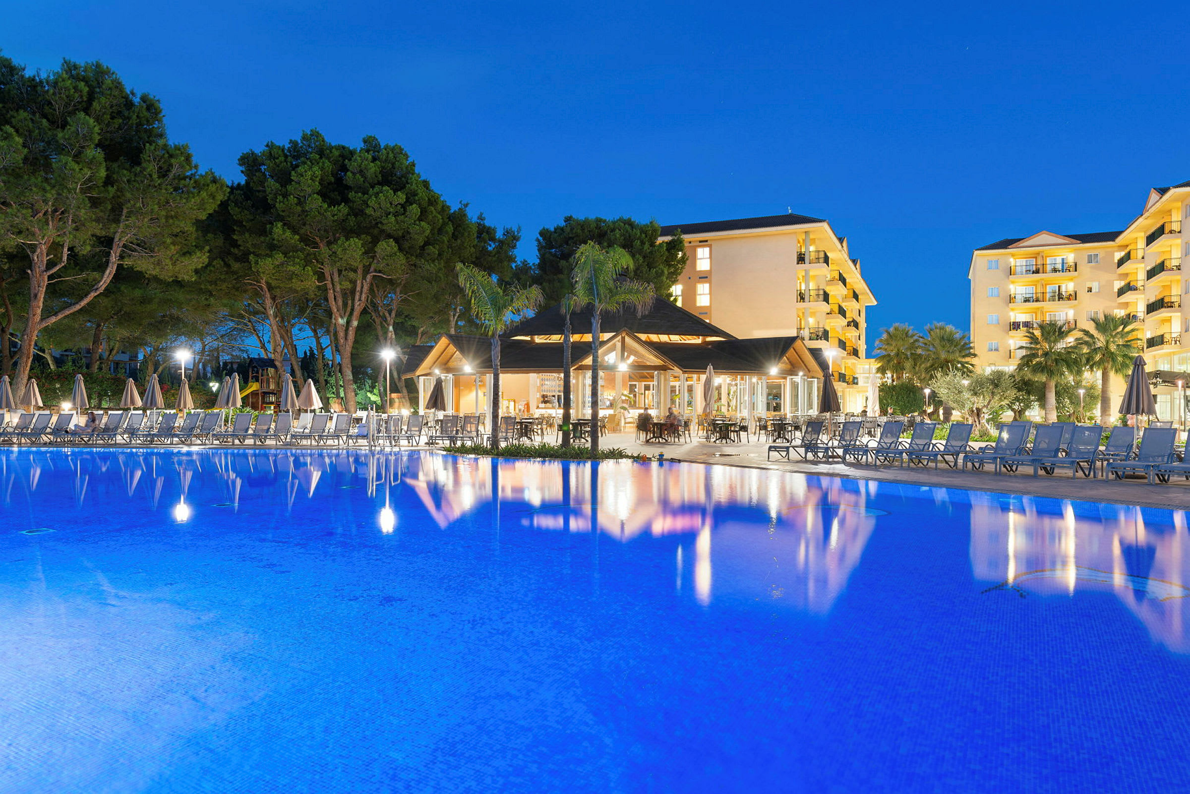 En oplyst hotelbygning mod en blå nattehimmel med en stor udendørs pool i forgrunden. (AI-genereret tekst)
