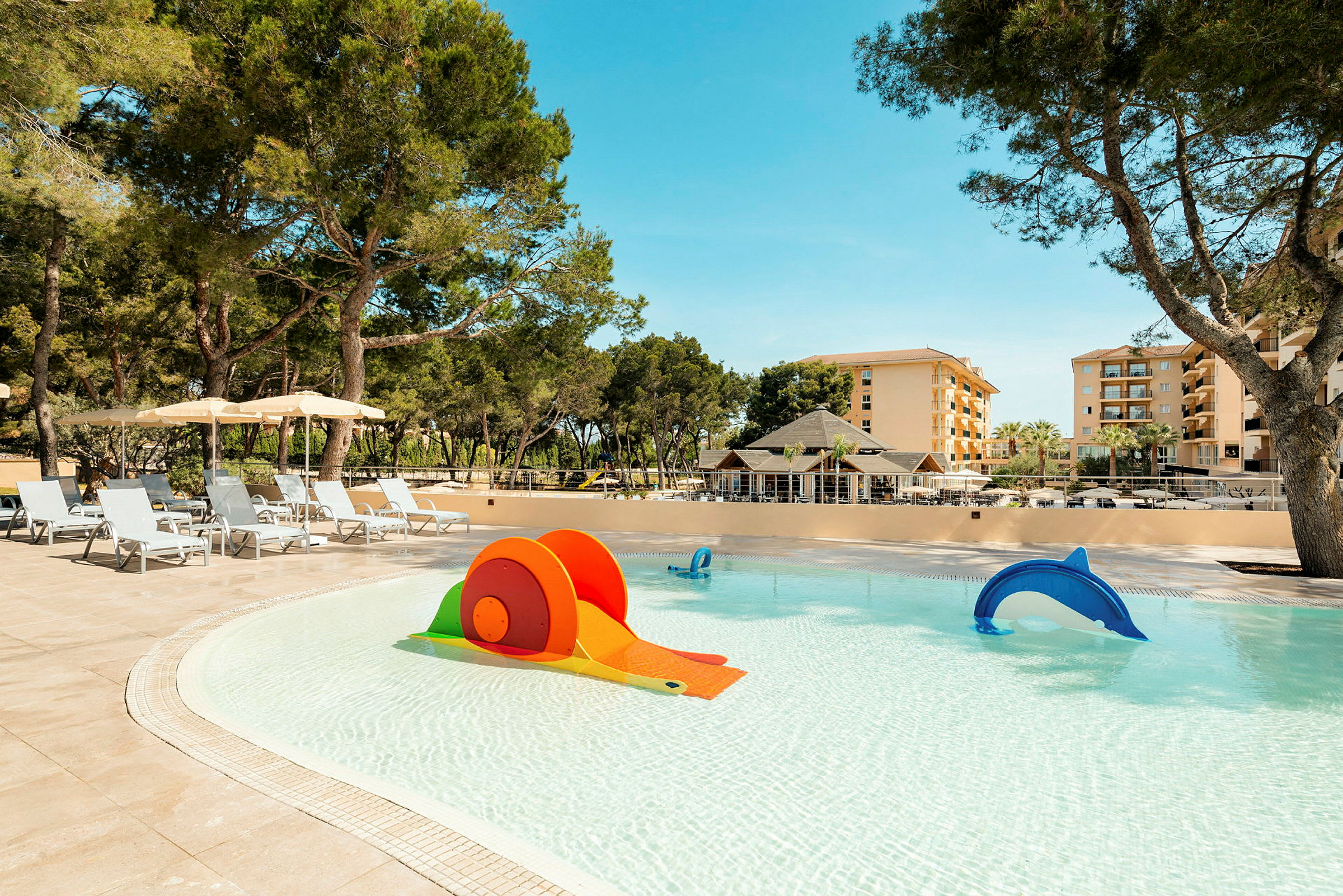 Billede af en swimmingpool med legetøj til børn, parasoller og omkringliggende vegetation, med bygninger i baggrunden. (AI-genereret tekst)