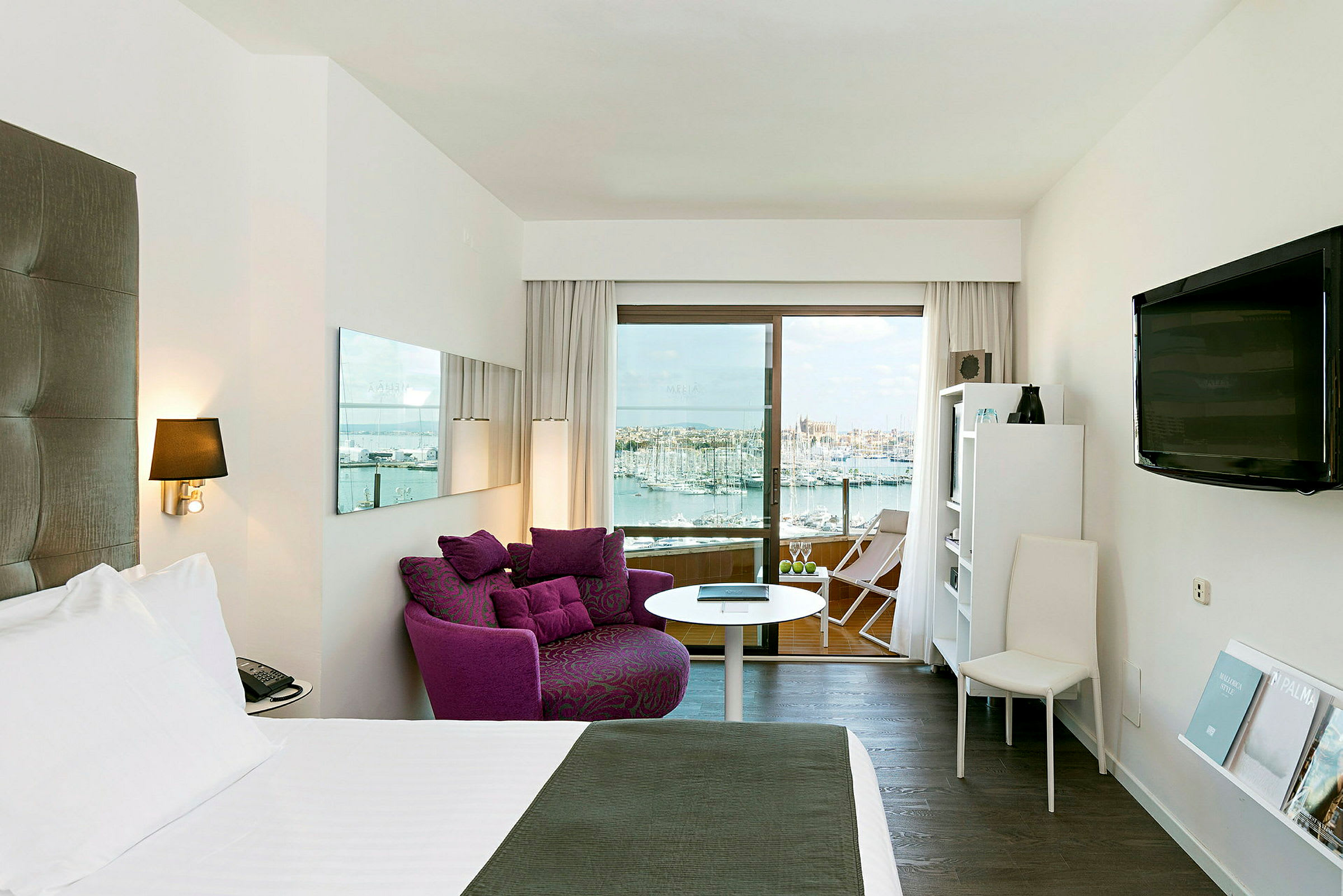Hotelværelse med en bred seng, lilla lænestol, et TV på væggen og en balkon med udsigt over havnen og byen. (AI-genereret tekst)