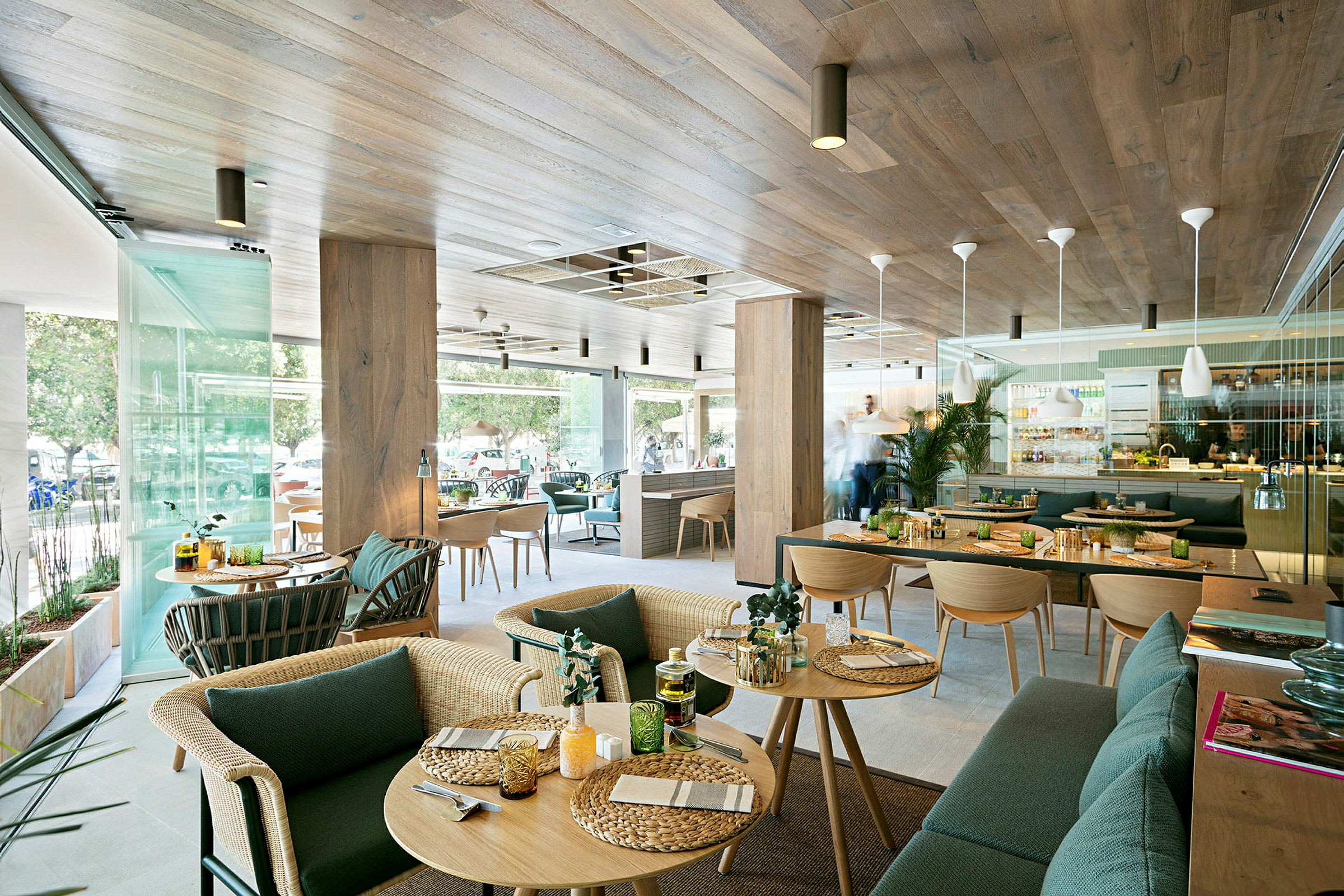 En moderne restaurant med træinteriør og store glasvinduer. Der er borde med serveret mad og interiør med grønne planter. (AI-genereret tekst)
