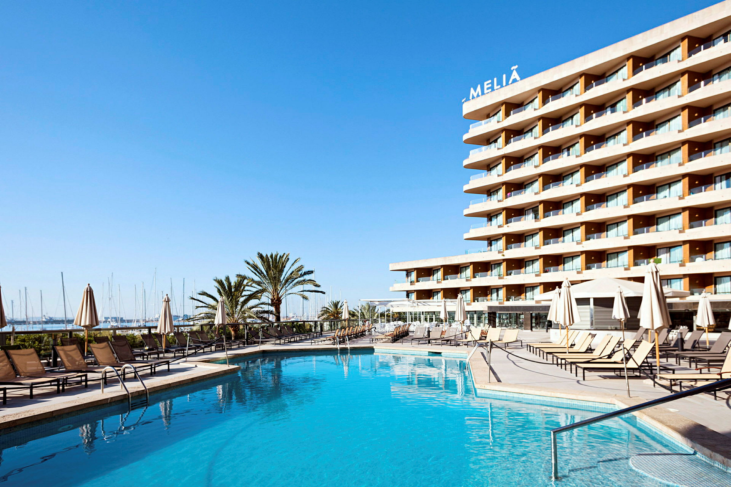 Et hotel med en swimmingpool i forgrunden, omgivet af liggestole og parasoller, med havet og en lystbådehavn i baggrunden under en klar blå himmel. (AI-genereret tekst)
