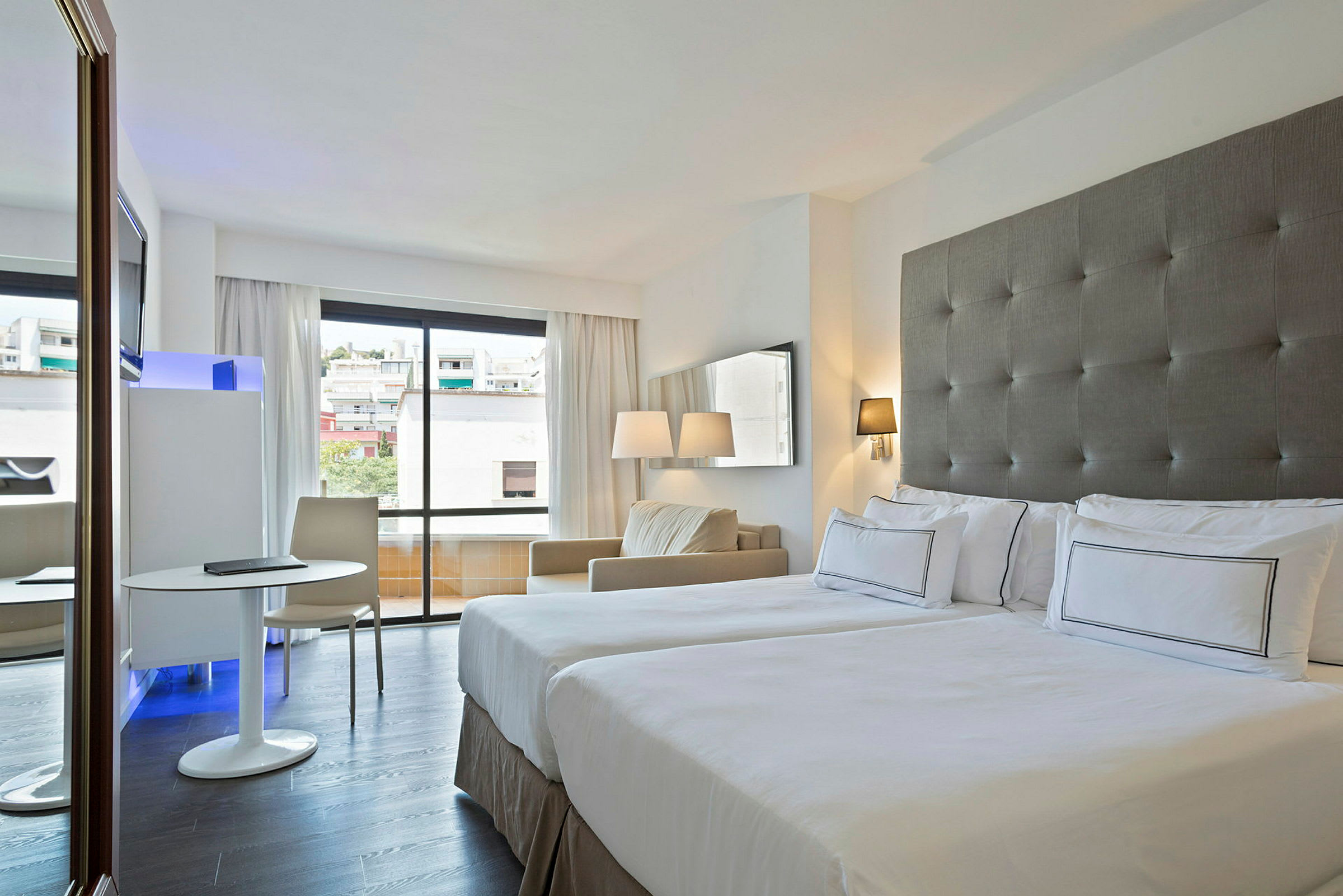 Hotelværelse med to senge, et vindue med udsigt til byen og et siddemiljø med bord og stol. (AI-genereret tekst)