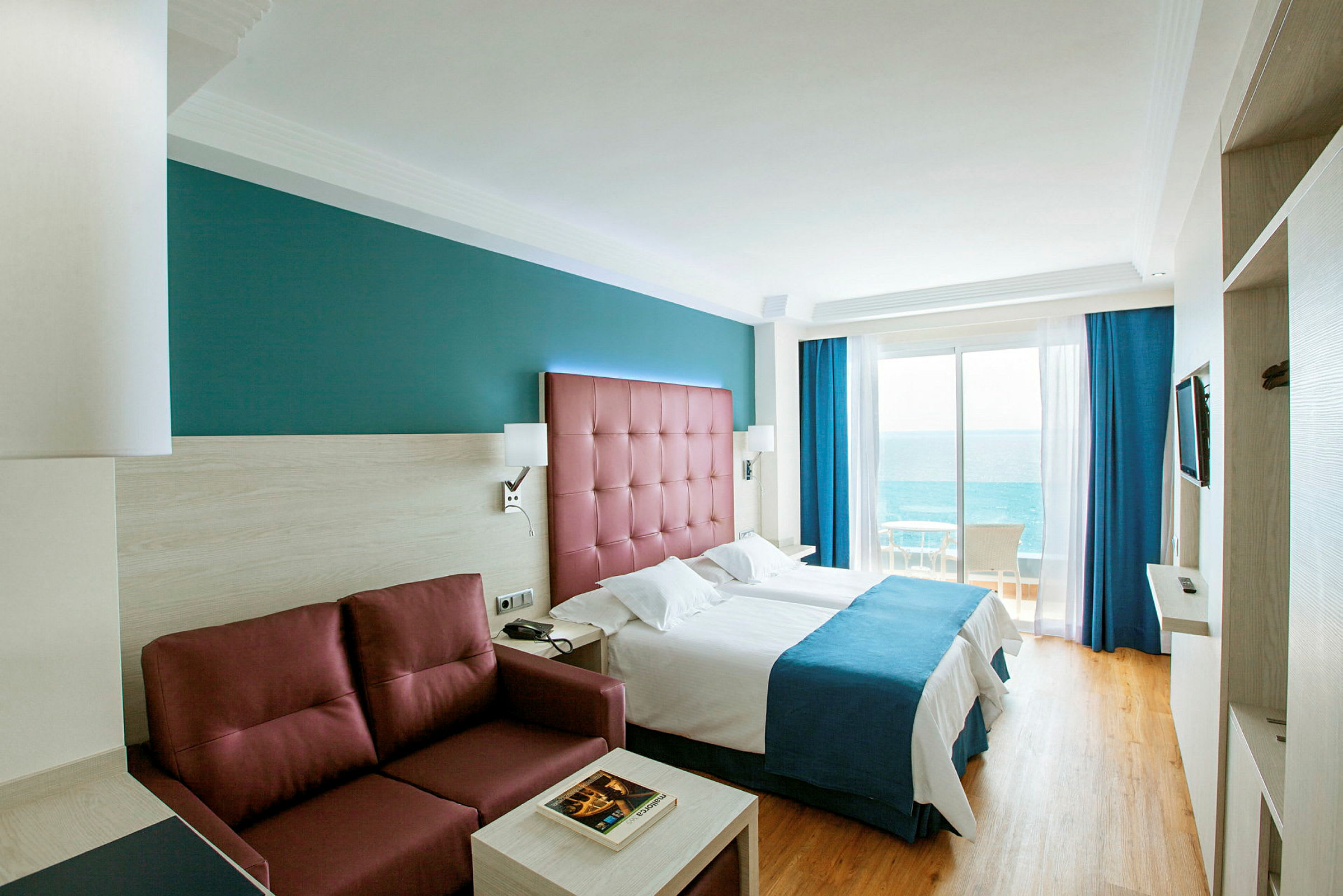 Hotelværelse med to enkeltsenge, en rød sofa og havudsigt fra altanen. (AI-genereret tekst)