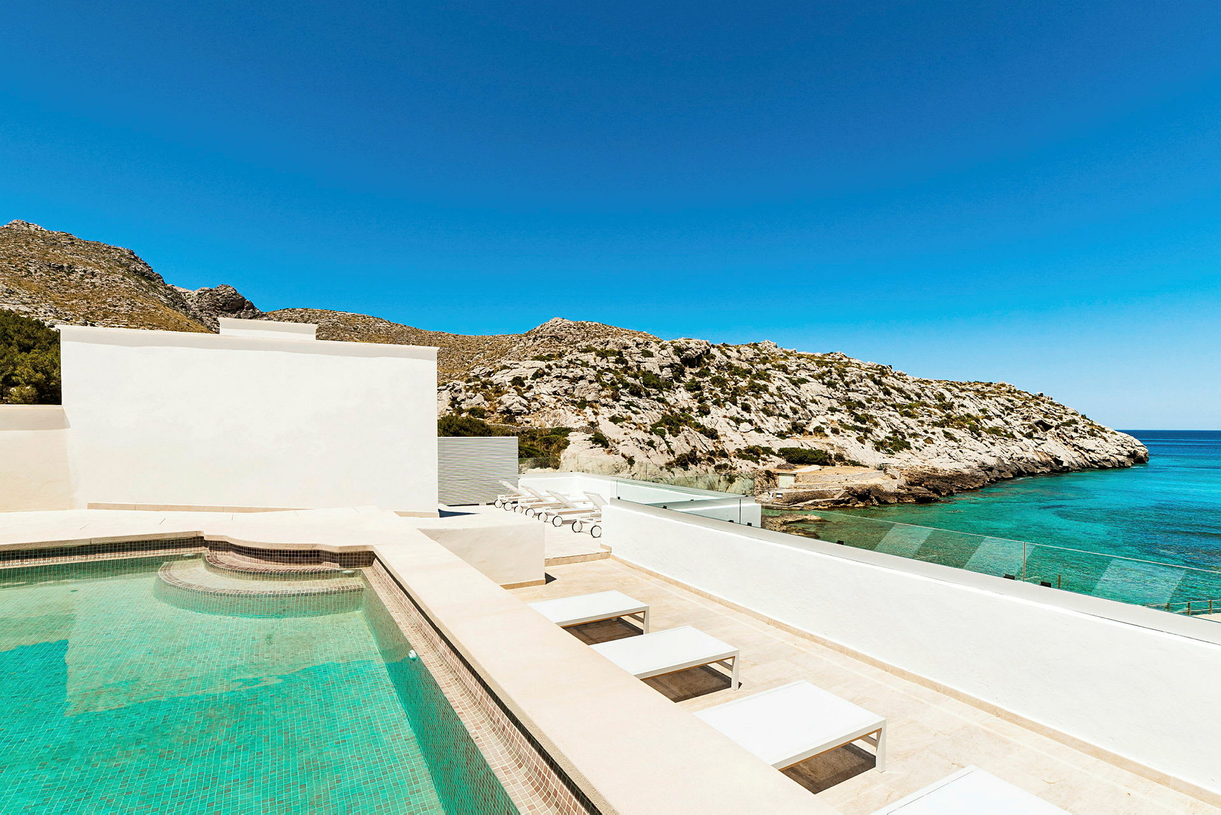 En udendørs pool ved et hus med udsigt over havet og klipper under en klar blå himmel. (AI-genereret tekst)
