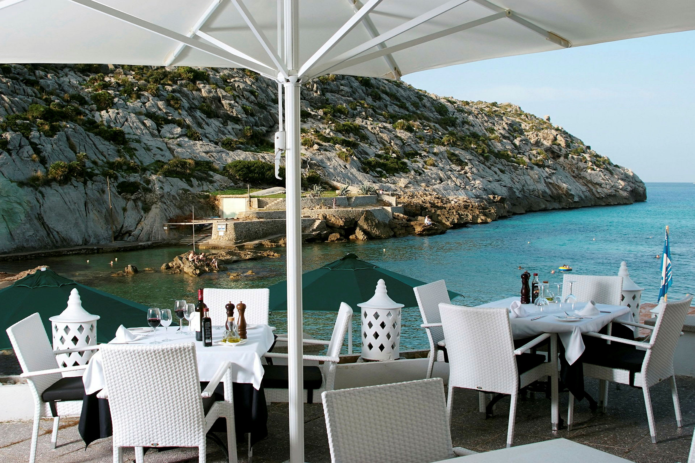 En udendørs restaurant med dækkede borde og hvide stole med udsigt til et klippefyldt kystlandskab og havet. (AI-genereret tekst)