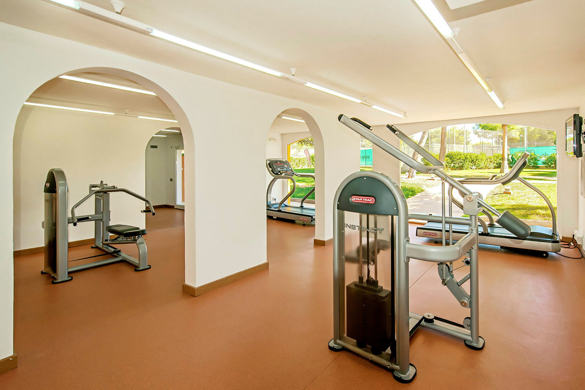 Et fitnesscenter med forskellige træningsmaskiner såsom løbebånd og styrketræningsudstyr. Rummet har store vinduer med udsigt til grønne områder. (AI-genereret tekst)