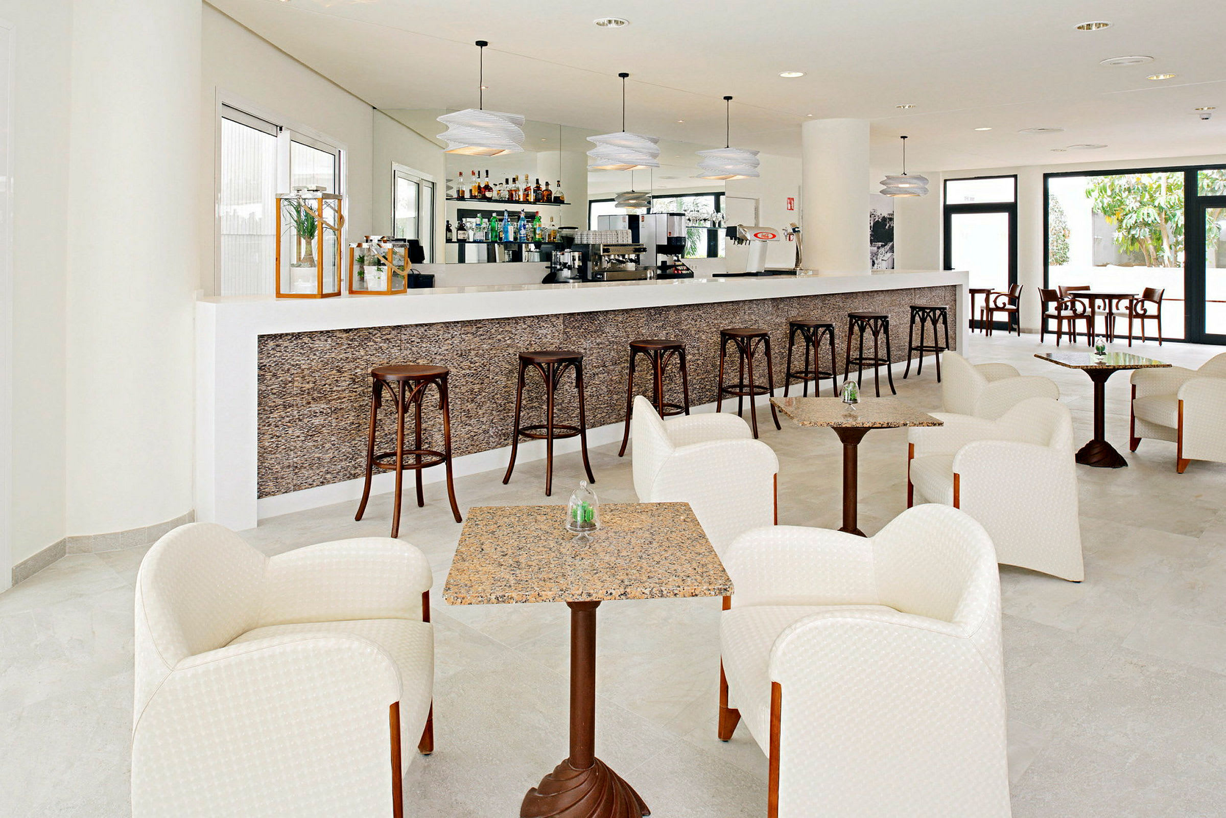Billede af en moderne hotelbar med en lang bardisk, høje barstole og siddegrupper med lænestole og små borde. (AI-genereret tekst)