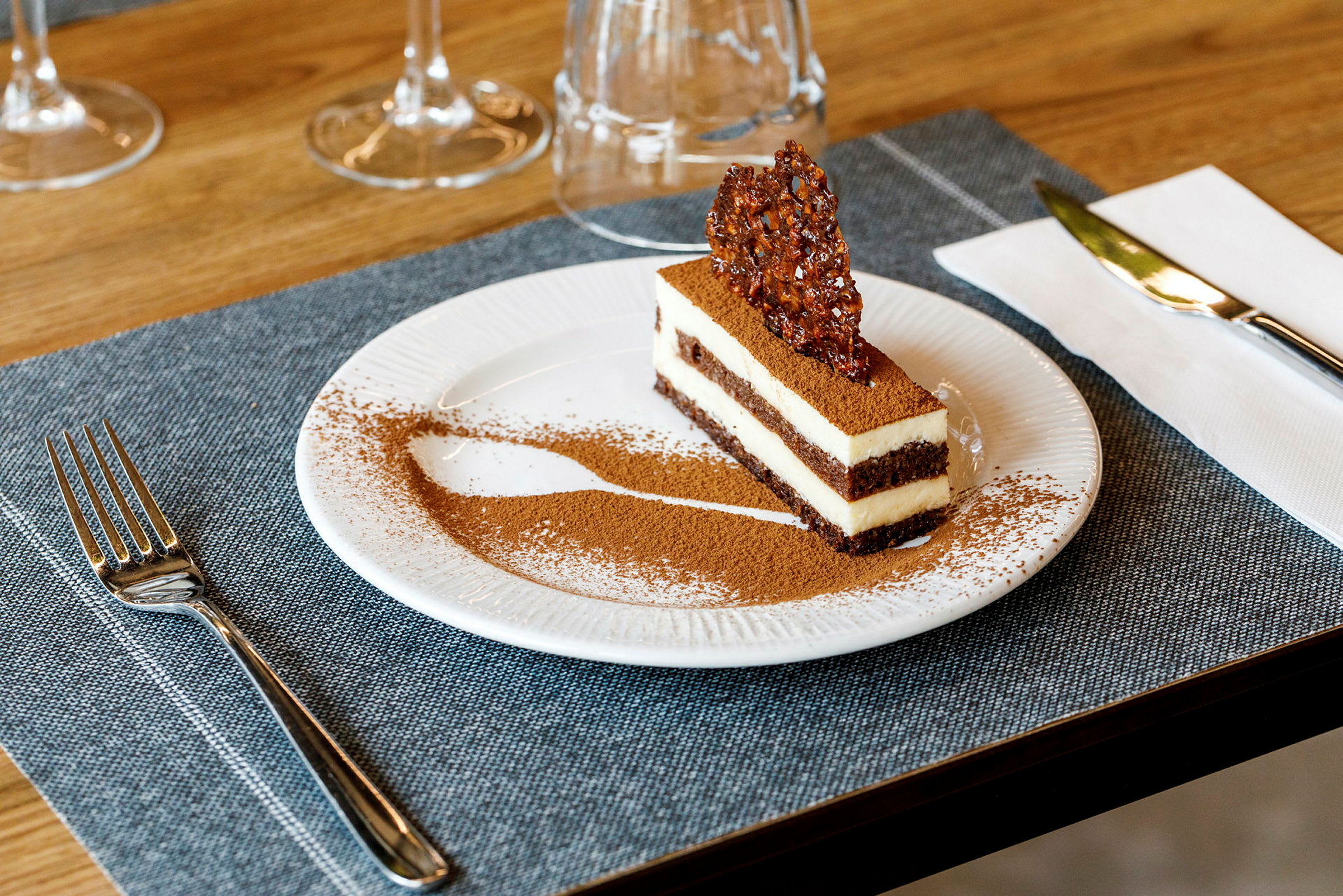 En dessert, sandsynligvis en tiramisu, serveret på en tallerken, dekoreret med kakaopulver og et sprødt stykke karamel på toppen. (AI-genereret tekst)