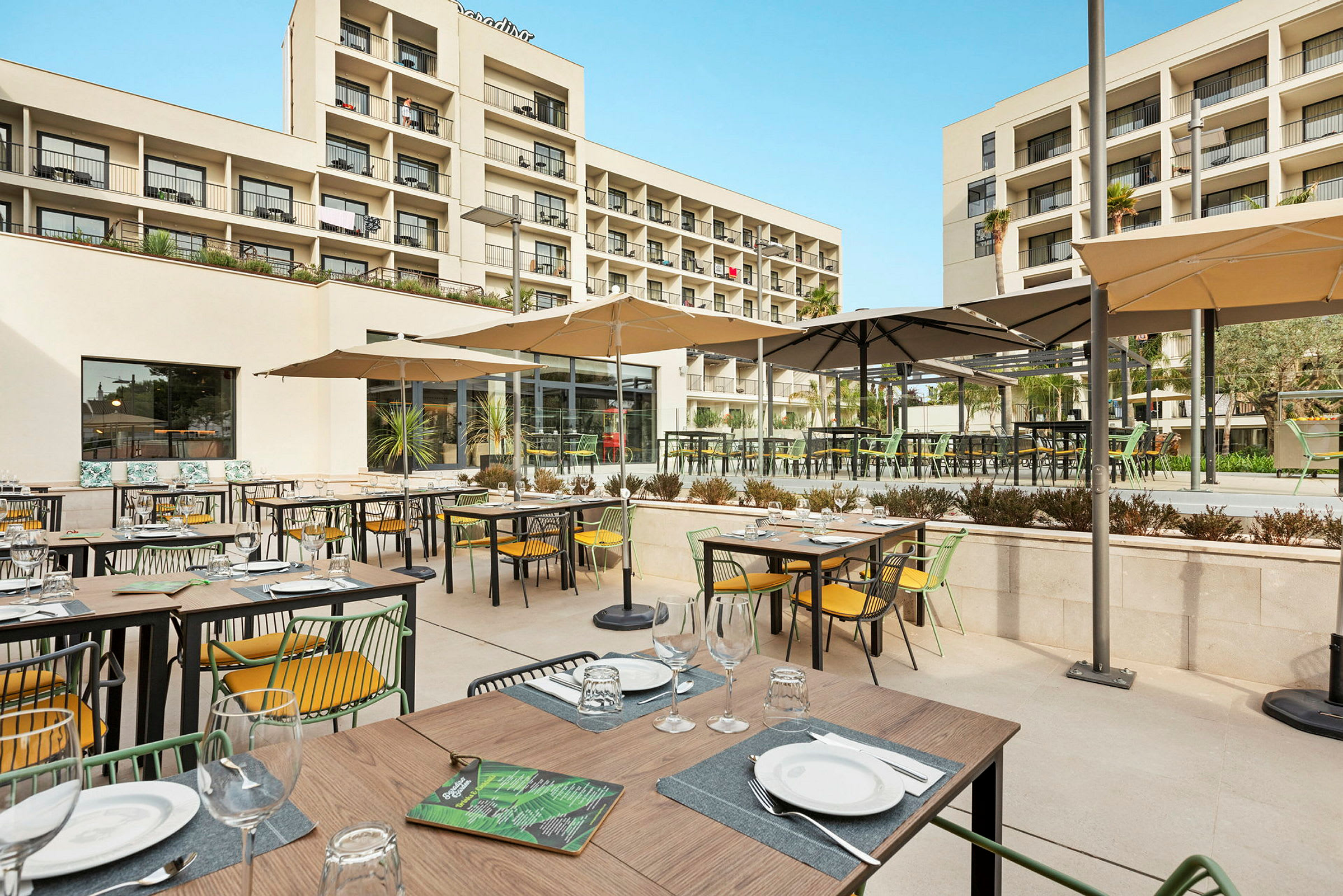 Billede af en udendørsrestaurant med opstillede borde og stole under parasoller samt hotelbygningen i baggrunden. (AI-genereret tekst)