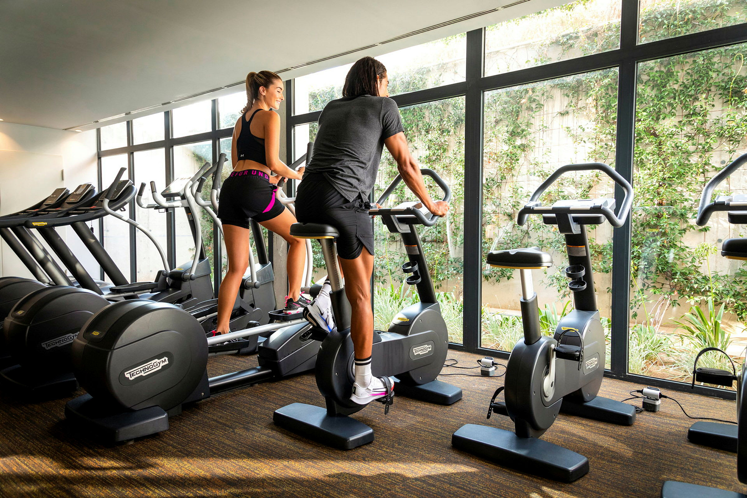 To personer træner på motionscykler i et fitnesscenter med store vinduer. (AI-genereret tekst)