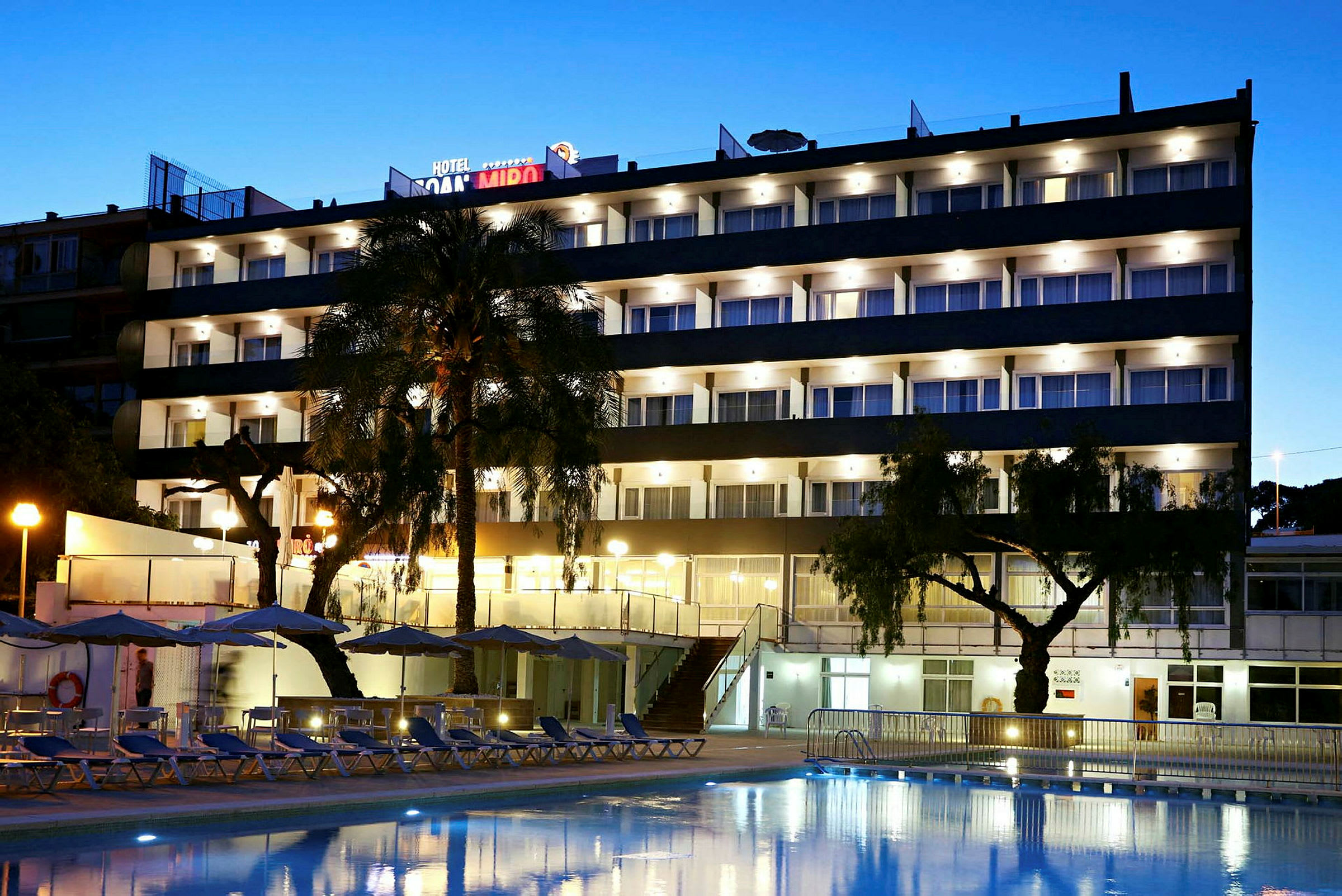 Et fleretagers hotel med altaner og oplyste vinduer, omgivet af træer, parasoller og en pool i forgrunden. (AI-genereret tekst)