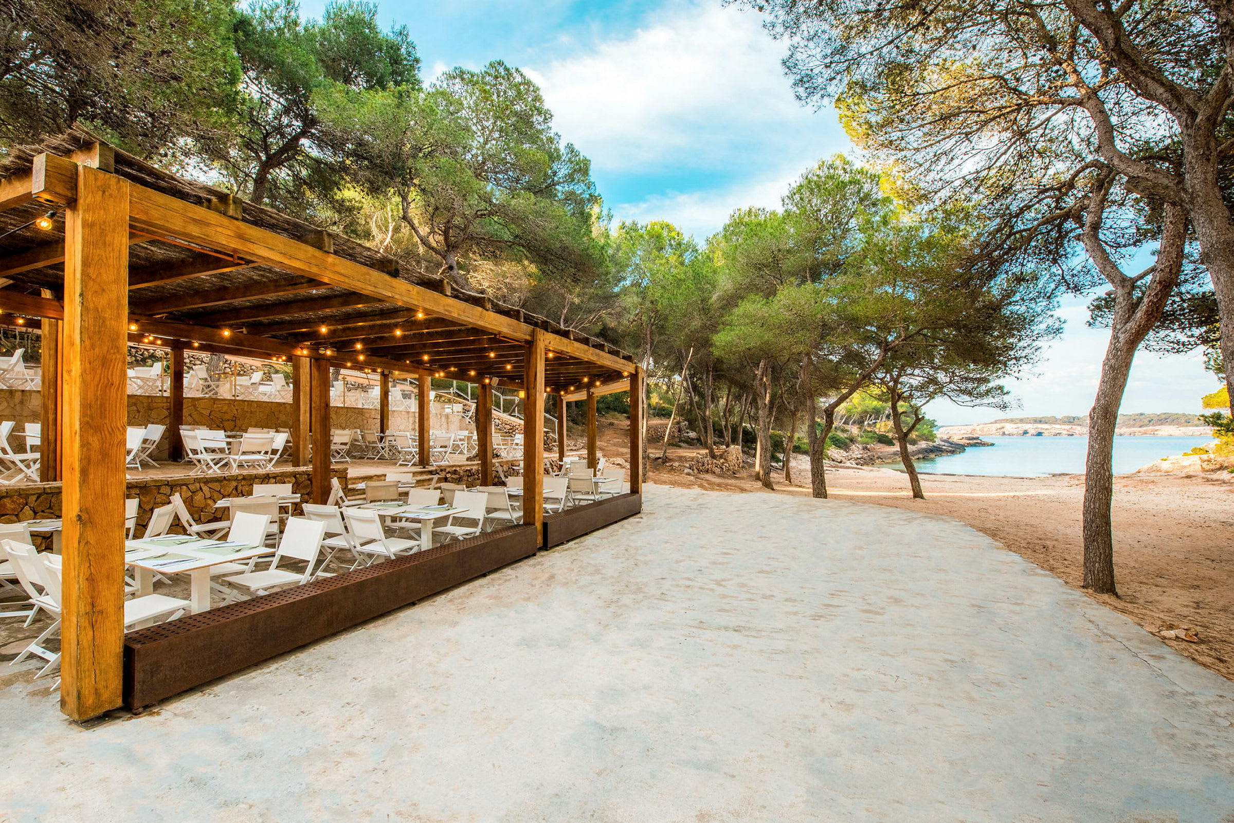 En strandrestaurant med hvide møbler og en træpergola tæt på sandstrand og træer. (AI-genereret tekst)