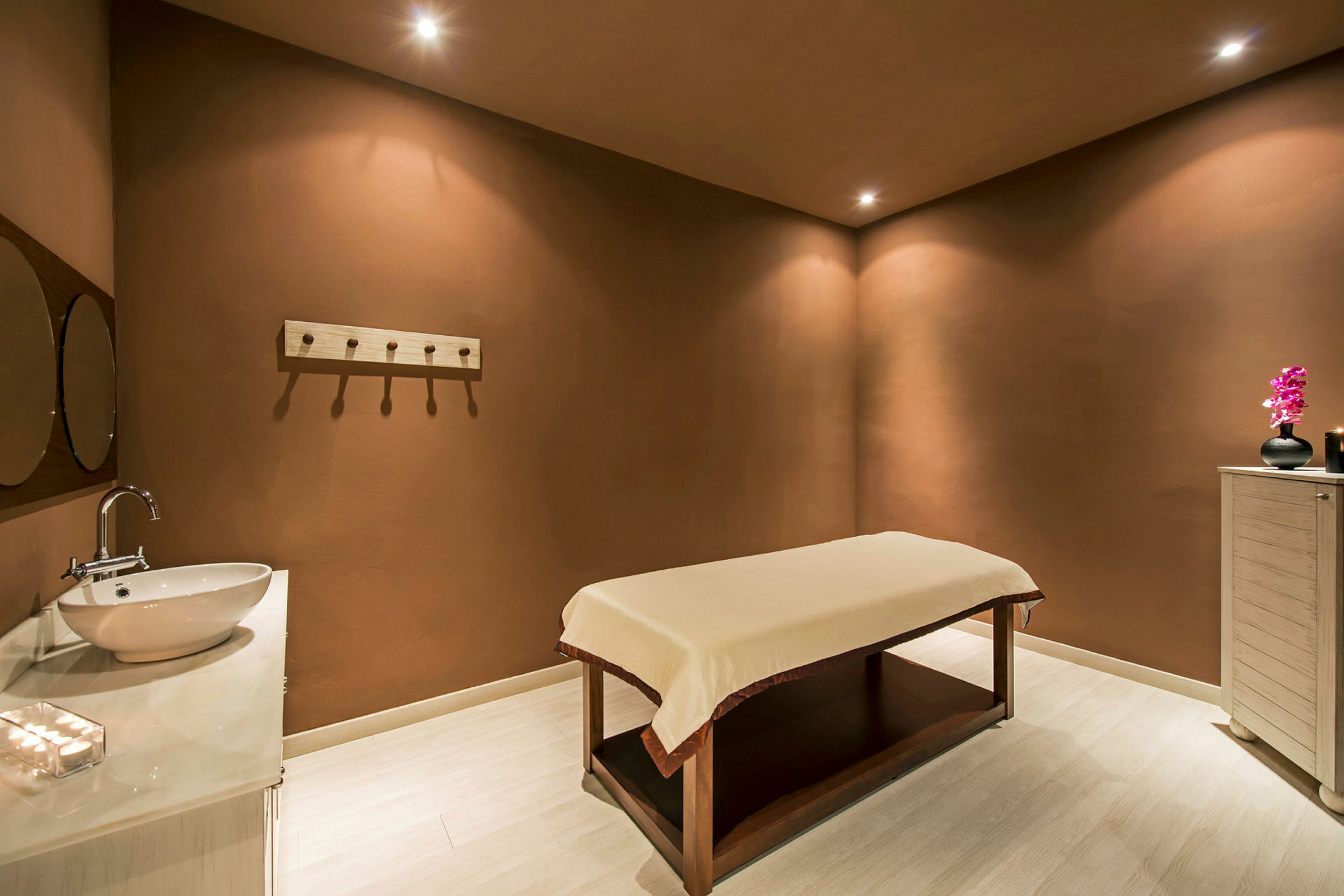 Et spa-rum med en massagebriks, en håndvask med spejl og et skab med en dekoration. (AI-genereret tekst)
