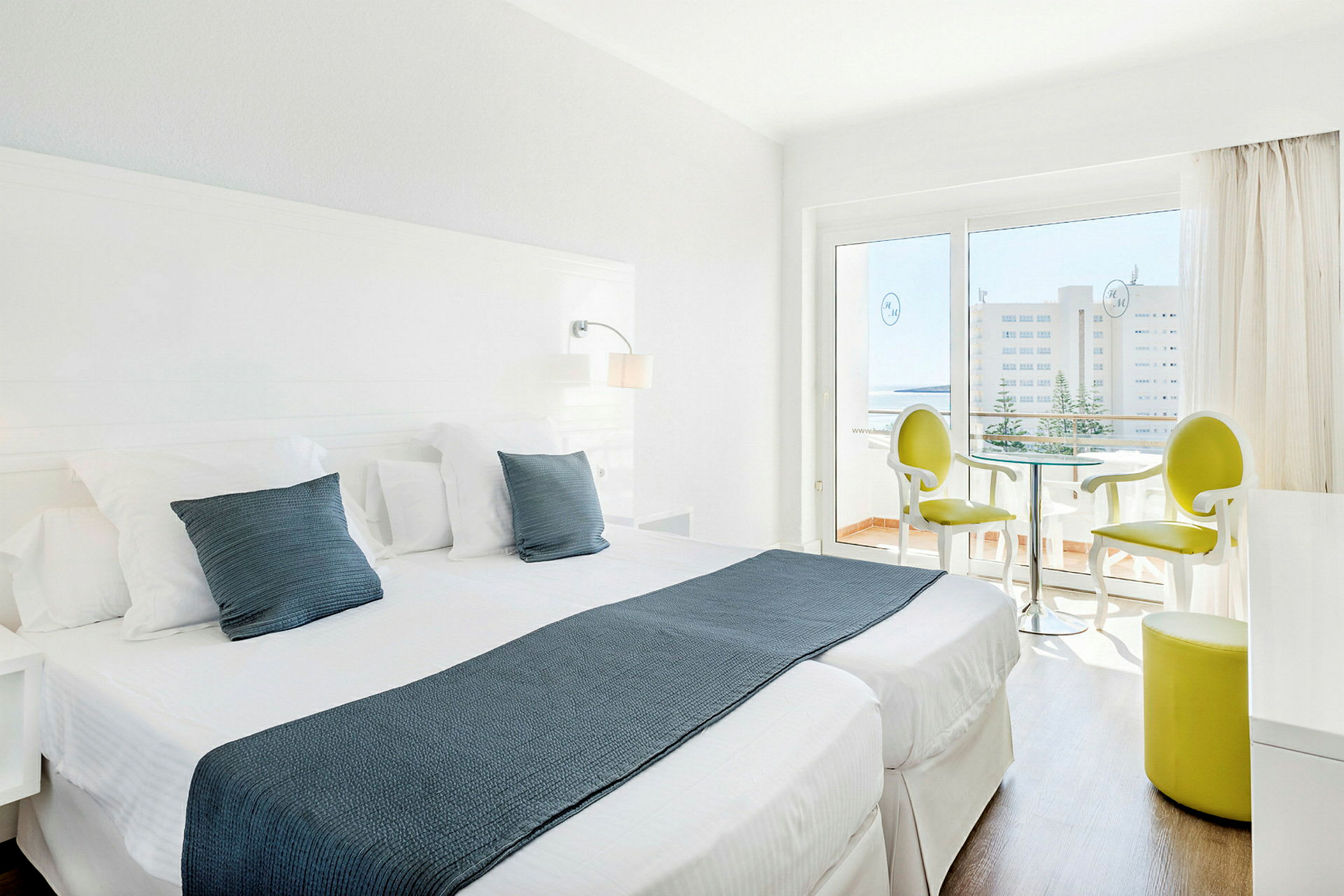 Et lyst hotelværelse med to enkeltsenge, hvide møbler og en siddegruppe nær en glasdør, der fører til en balkon. (AI-genereret tekst)