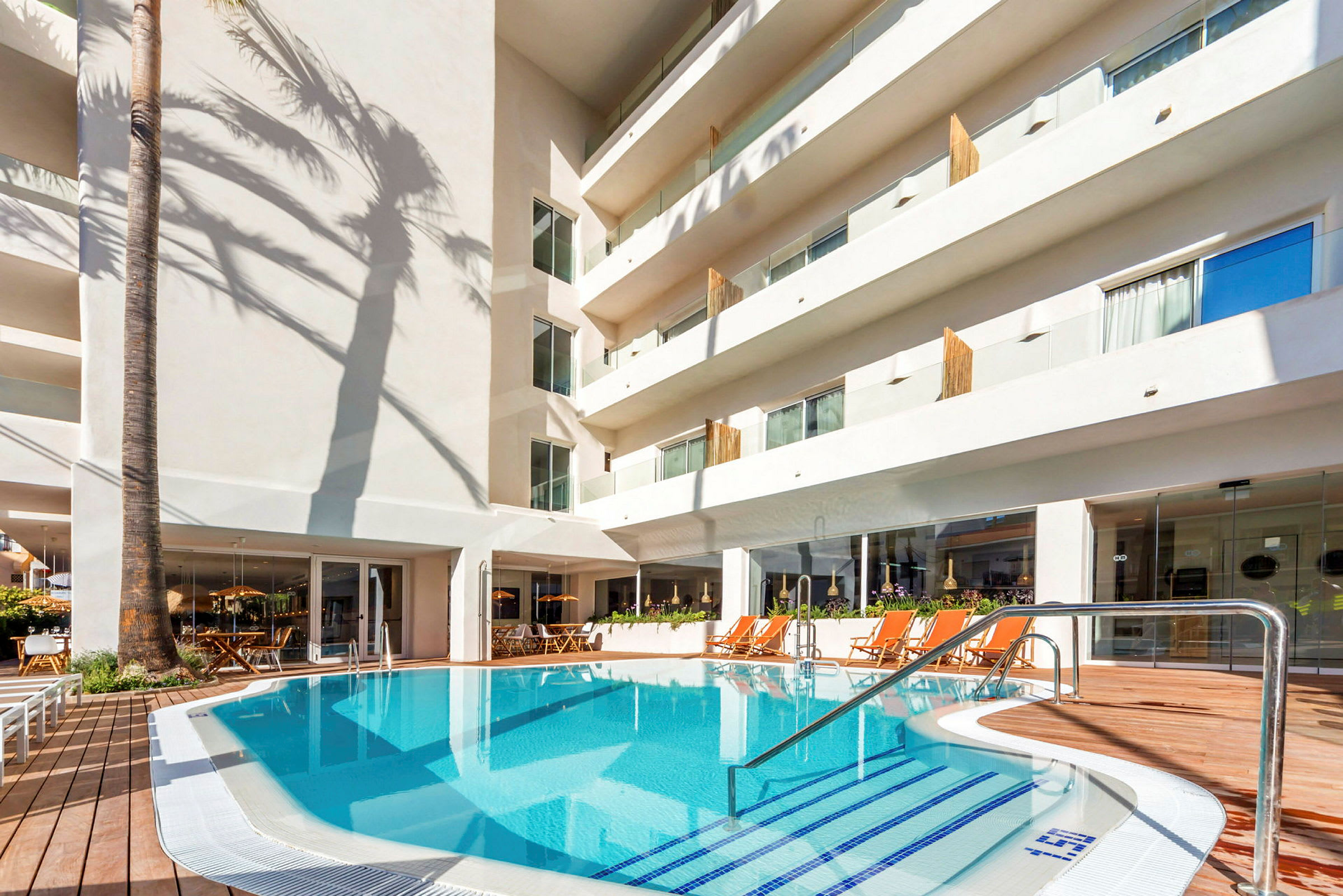 En hotelpool omgivet af liggestole og en hvid bygning. (AI-genereret tekst)