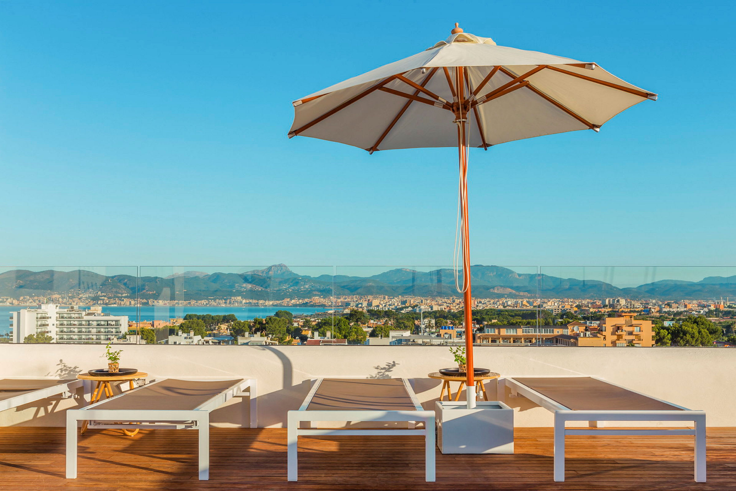 En solrig tagterrasse med liggestole, en parasol og udsigt over en by og bjerge i baggrunden. (AI-genereret tekst)