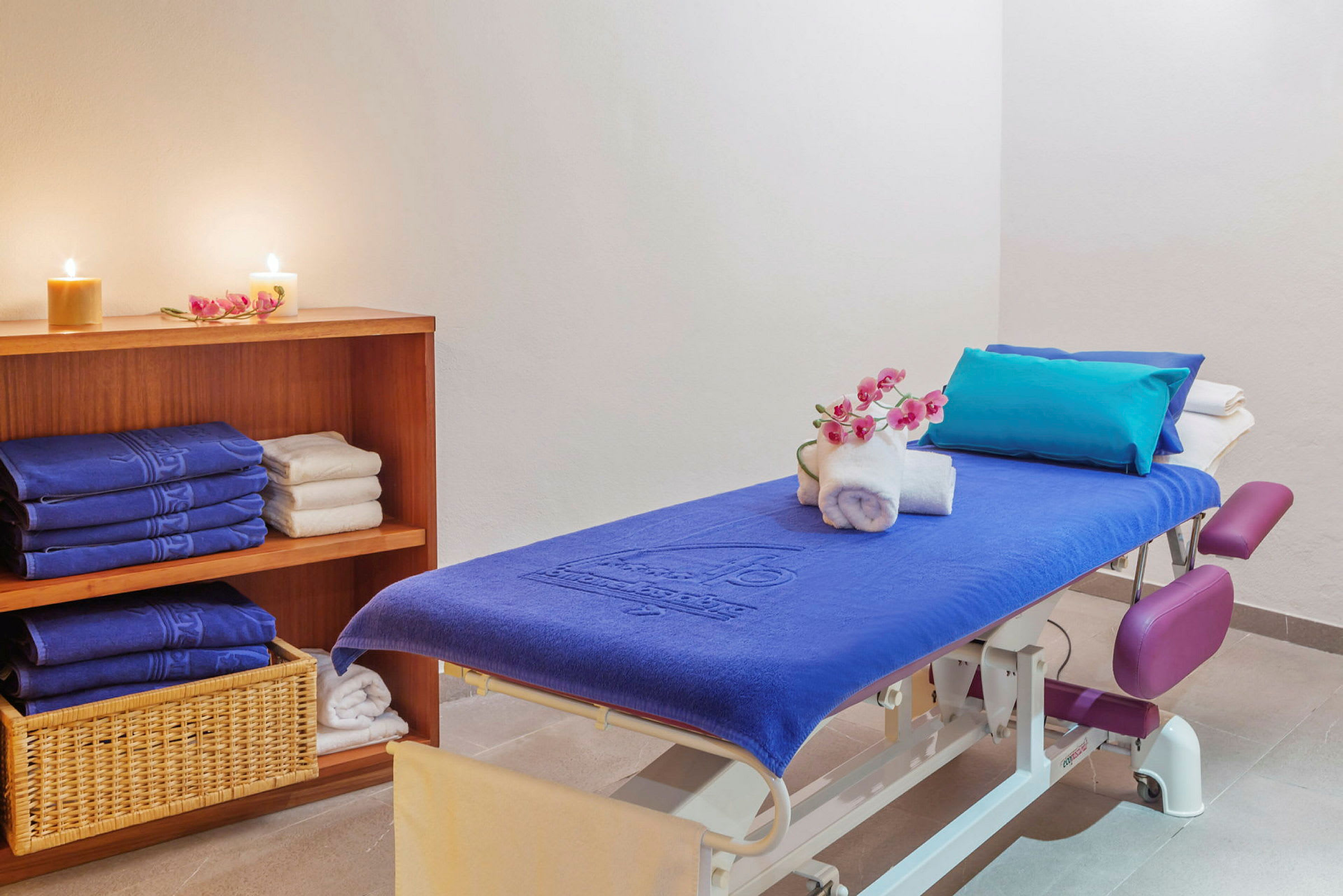 Et massagerum med en massagebriks dækket med et blåt håndklæde, dekoreret med hvide håndklæder og en blomsterkvist. I baggrunden er en hylde med foldede håndklæder og tændte lys. (AI-genereret tekst)