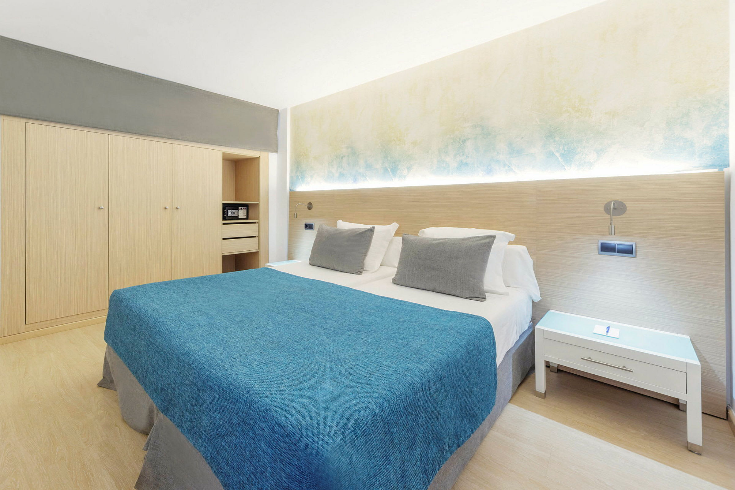 Hotelværelse med dobbeltseng, blåt sengetæppe, hvide puder og minimalistisk indretning. (AI-genereret tekst)