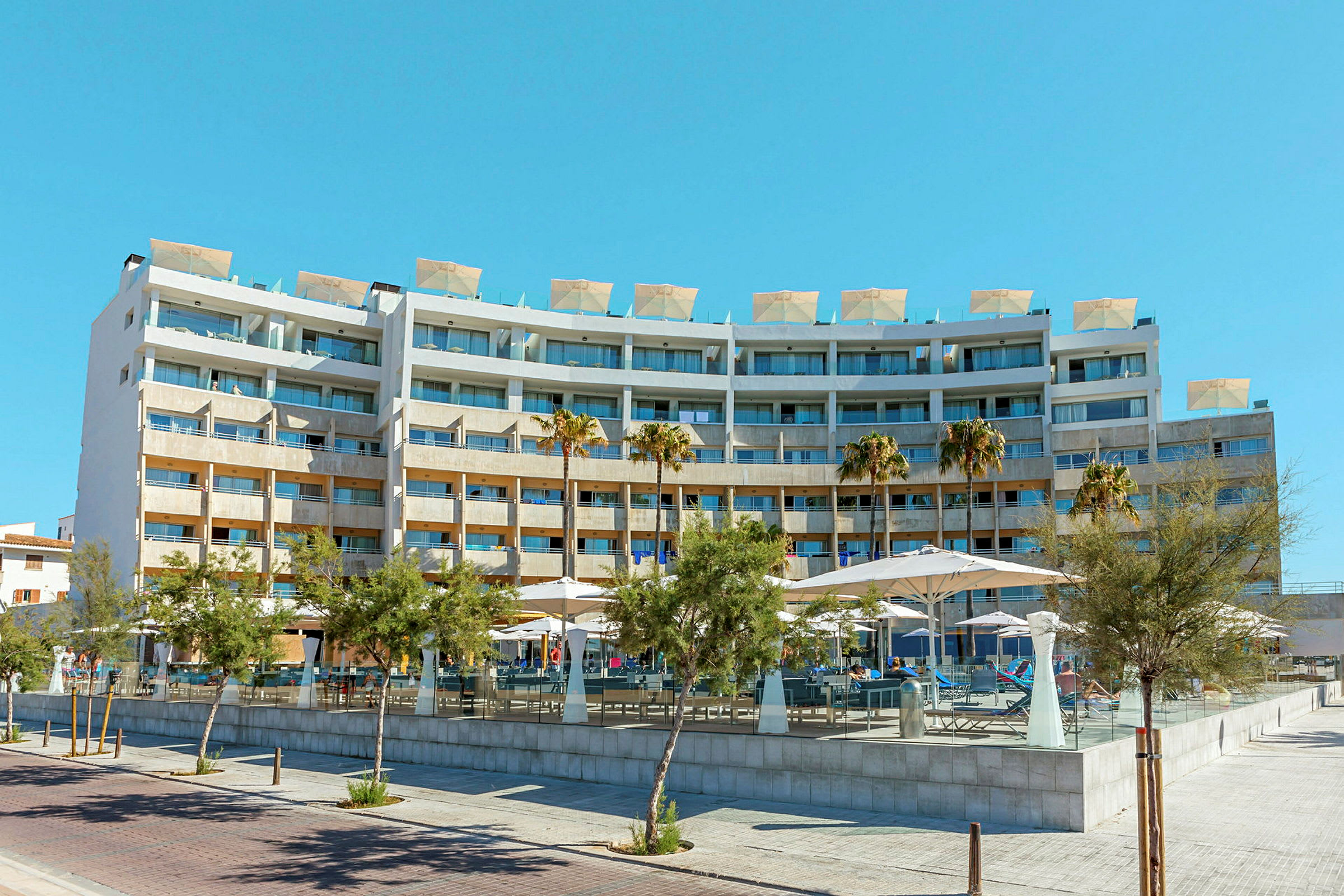 Et større hotelkompleks med flere etager og altaner. Foran bygningen er der nogle træer og parasoller. (AI-genereret tekst)
