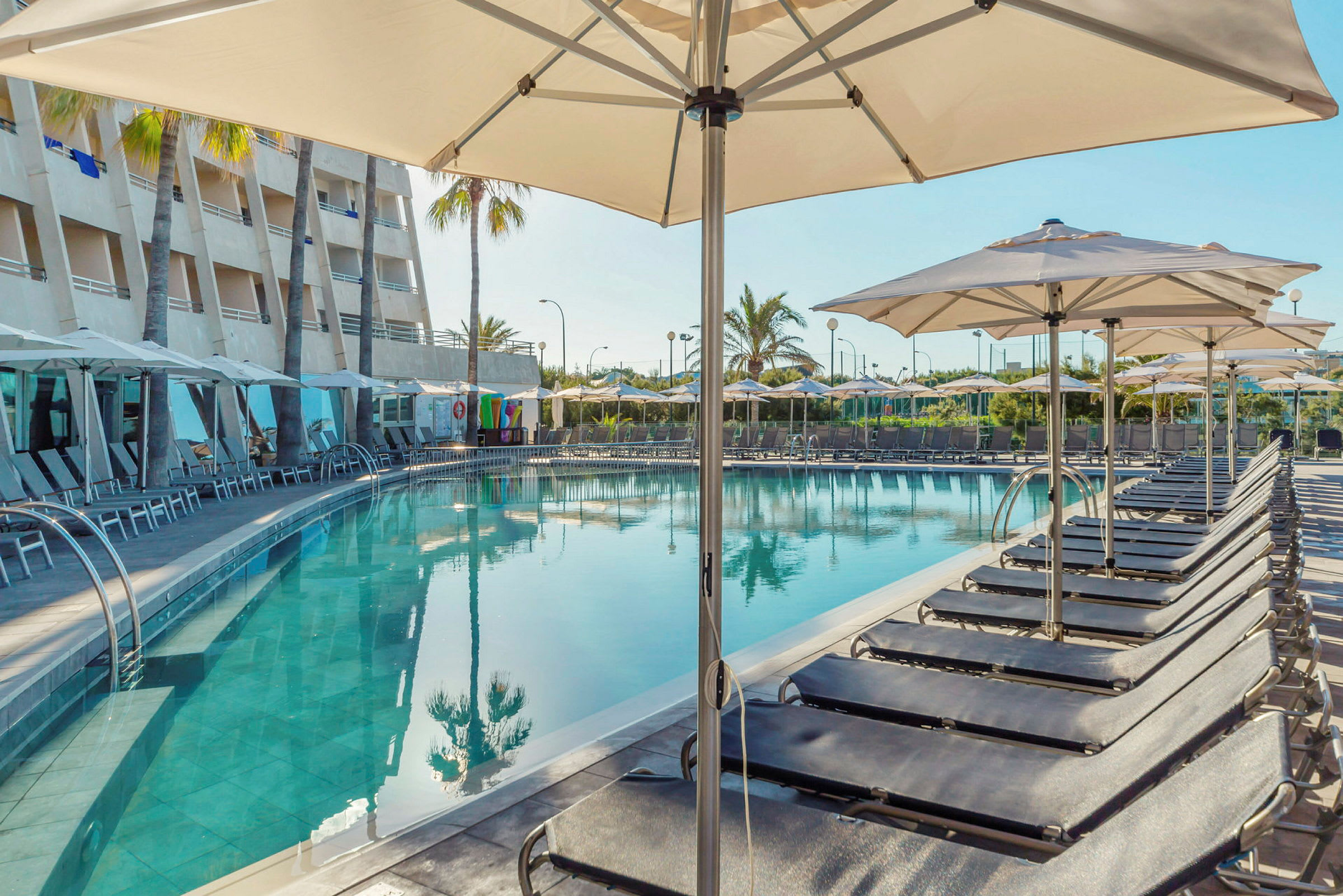 Hotelpool med liggestole og parasoller ved siden, samt hotelbygning og palmer i baggrunden. (AI-genereret tekst)