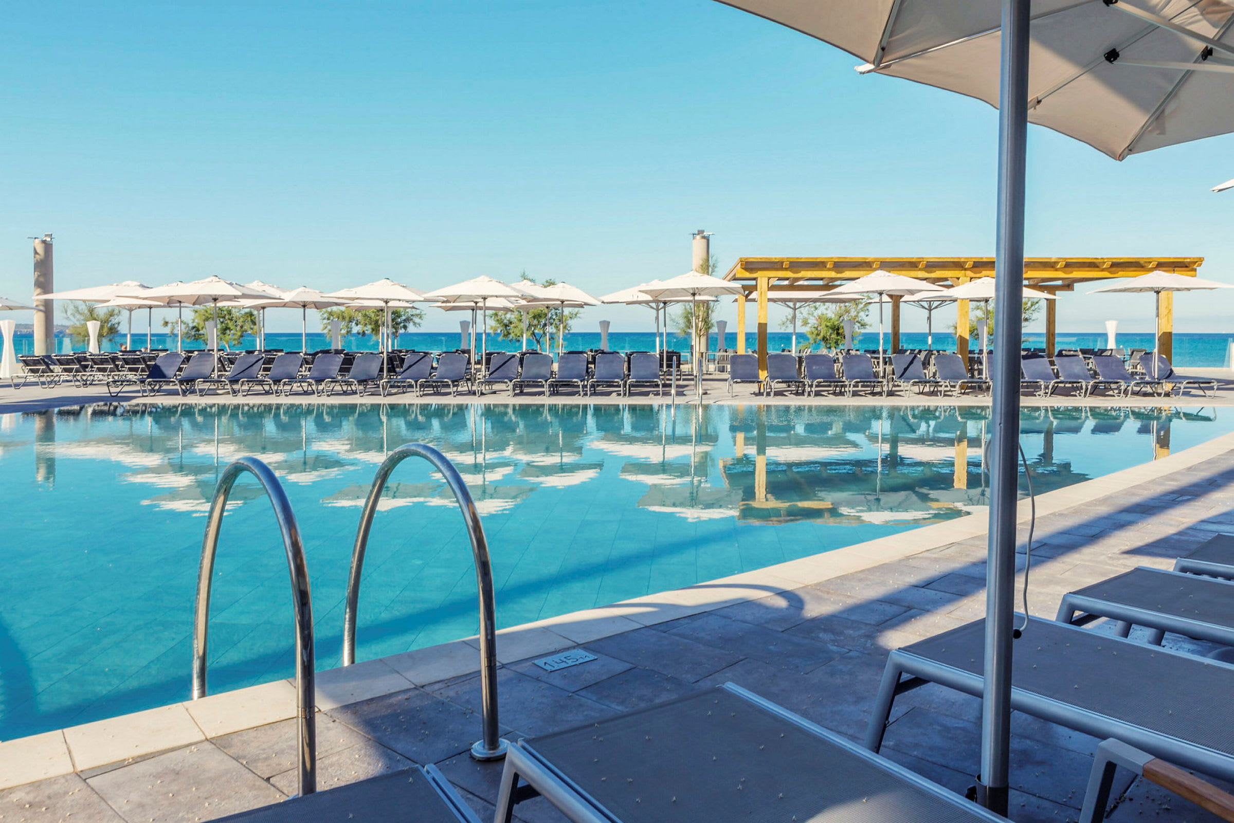 En pool med liggestole og parasoller i en udendørs omgivelser tæt på havet. (AI-genereret tekst)