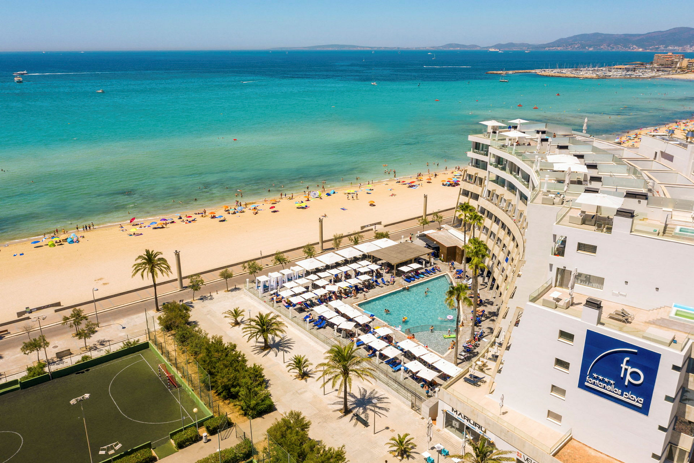 Udsigt over en strand med klart turkisblåt hav og en hotelresort med pool i forgrunden. (AI-genereret tekst)