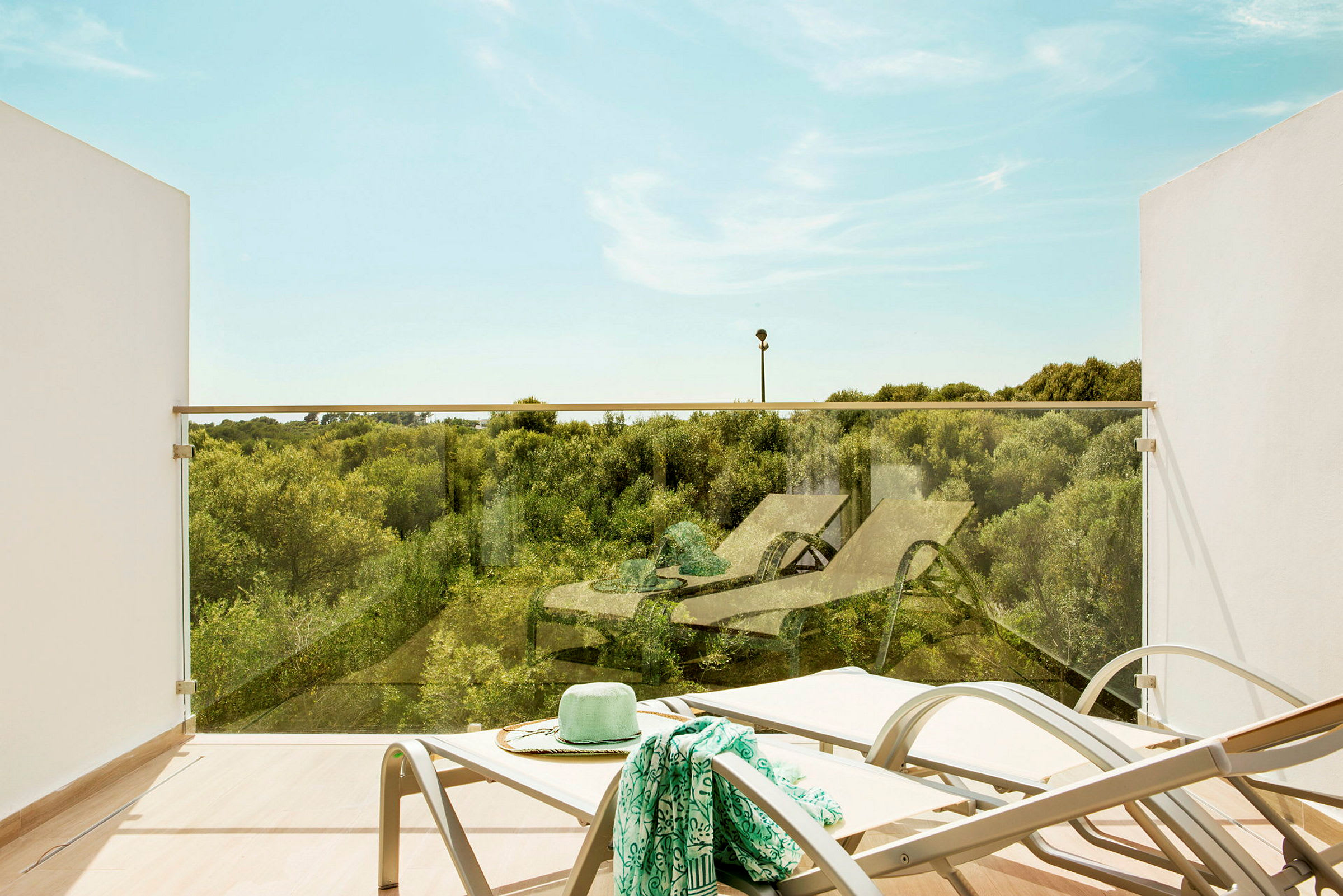 En terrasse med udsigt over grønne områder, liggestole og et glasrækværk. (AI-genereret tekst)