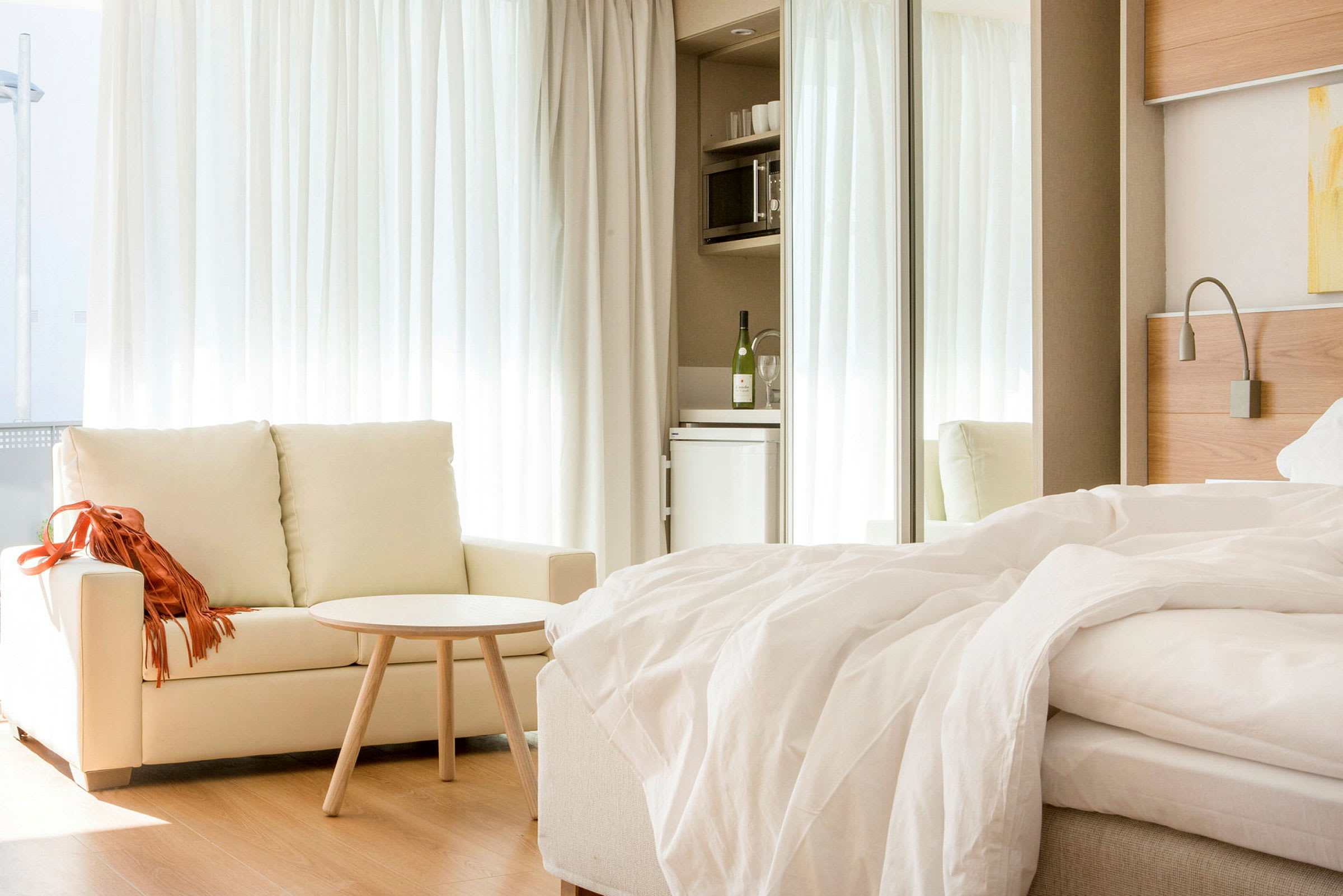 Et lyst hotelværelse med en sofa, et lille bord og en redt seng. I baggrunden er der et tekøkken med køleskab. (AI-genereret tekst)