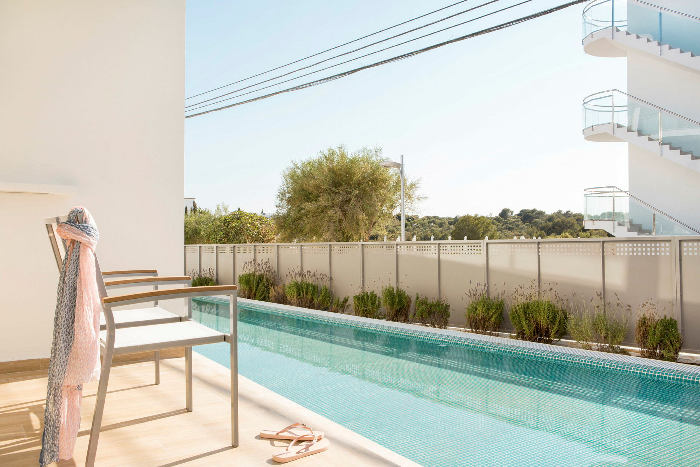En swimmingpool med klart blåt vand omgivet af et hegn og grønne planter, med to stole på en solterrasse og en trappe i baggrunden. (AI-genereret tekst)