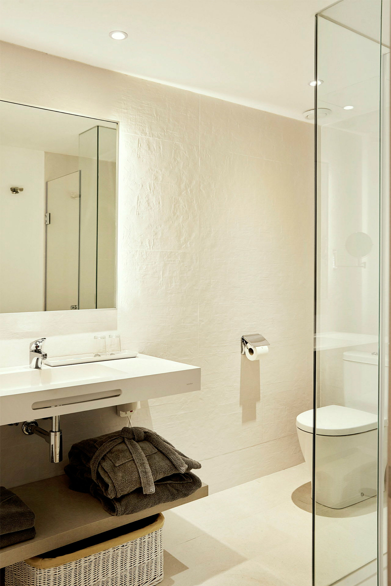 Et moderne badeværelse med hvid porcelænsvask, toilet og bruser bag glasdør. Der er håndklæder og kurve organiseret i rummet. (AI-genereret tekst)