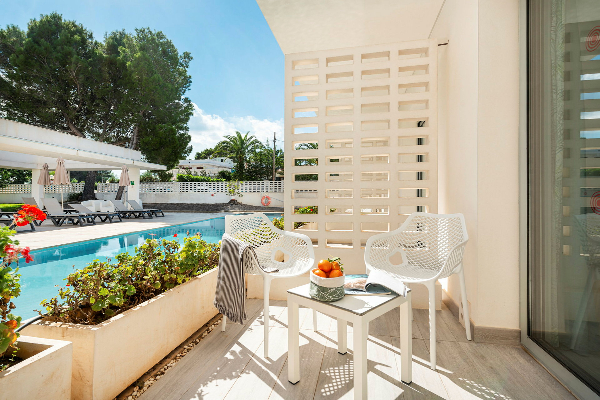Billede af en balkon med to hvide stole og et bord med appelsiner, med udsigt til en pool og have. (AI-genereret tekst)