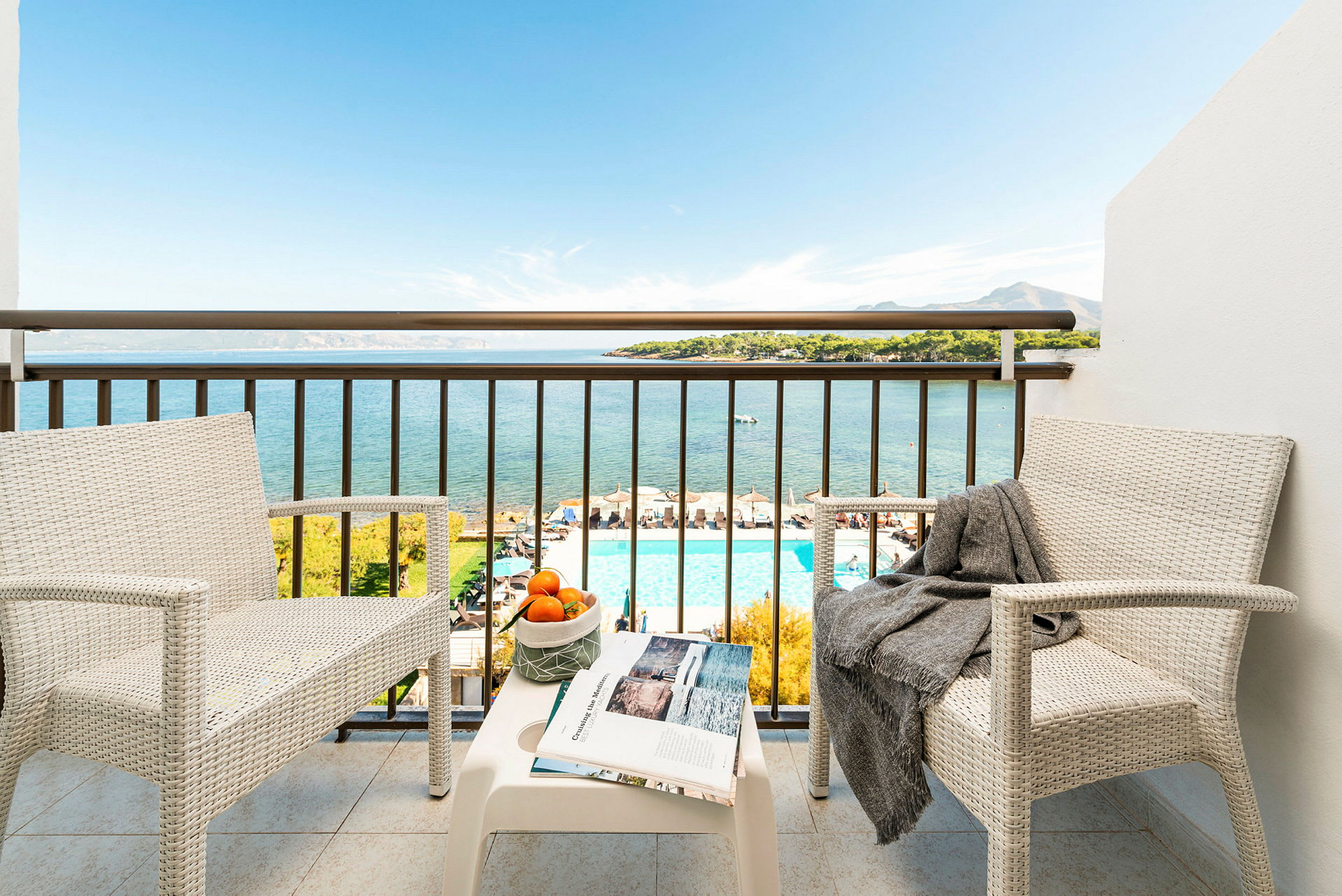 To hvide stole med et bord på en balkon, der giver udsigt over havet, en pool og landskabet. (AI-genereret tekst)