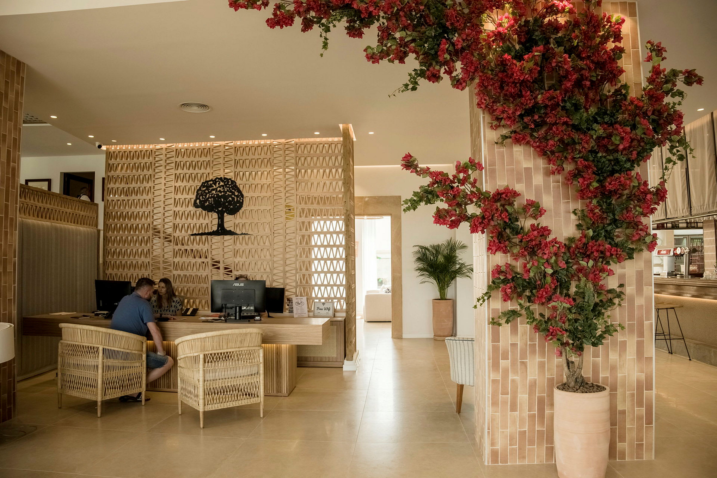 Et receptionsområde med træmøbler og en vægdekoration af en silhouet af et træ. Røde blomster pryder en plante i en beige krukke til højre. (AI-genereret tekst)
