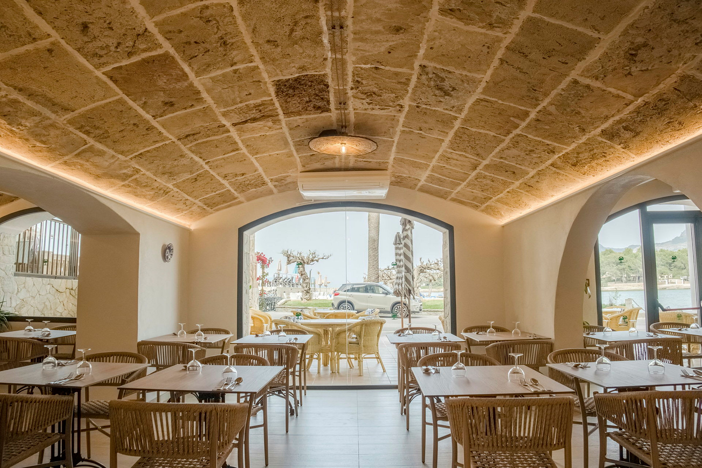 Interiør af en restaurant med stenvægge og hvælvet loft, borde og stole i træ samt store vinduer med udsigt til udeserveringsområde. (AI-genereret tekst)