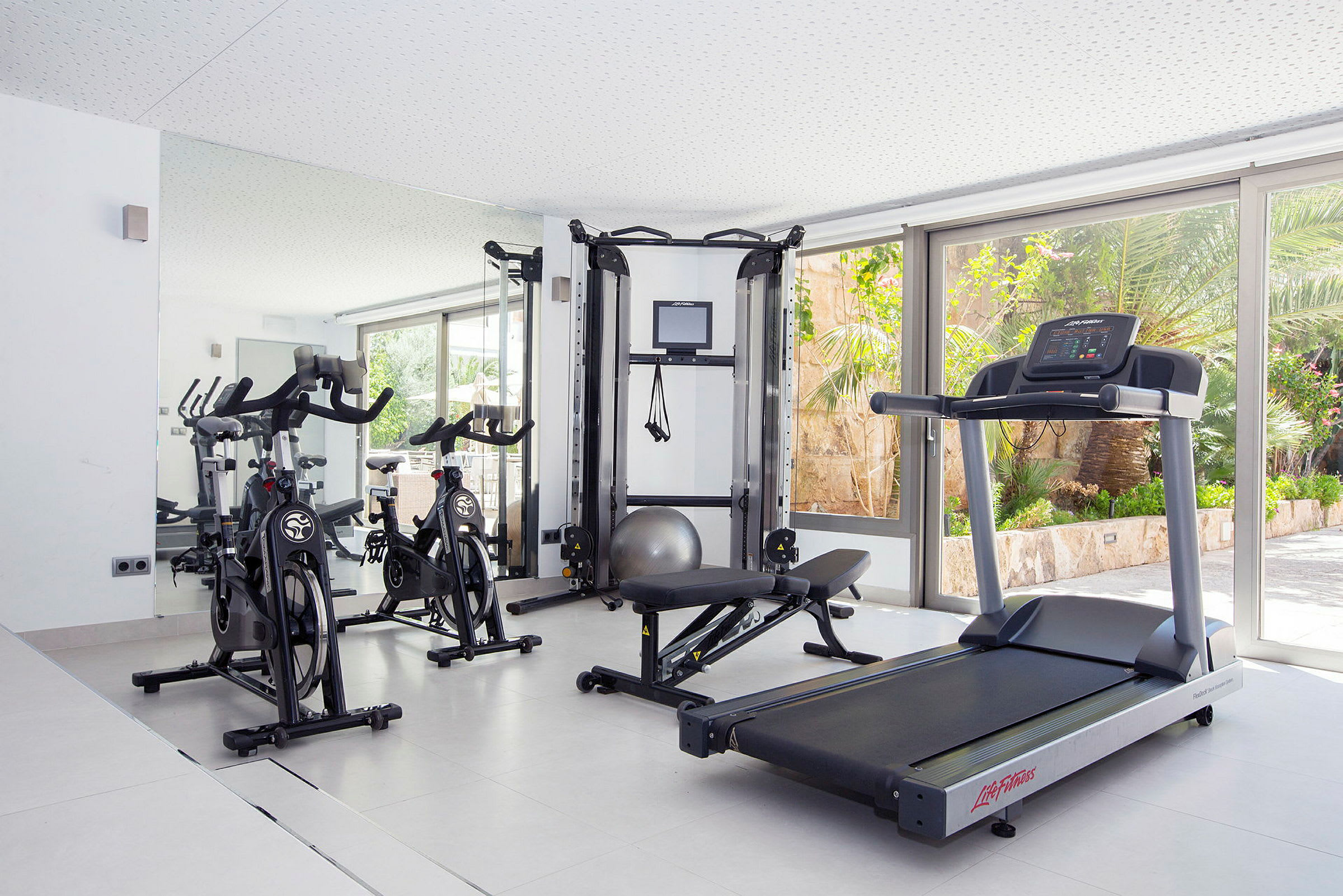 Billede af et fitnesscenter med træningsudstyr som motionscykler, løbebånd og styrketræningsmaskine i et lyst rum med store vinduer. (AI-genereret tekst)
