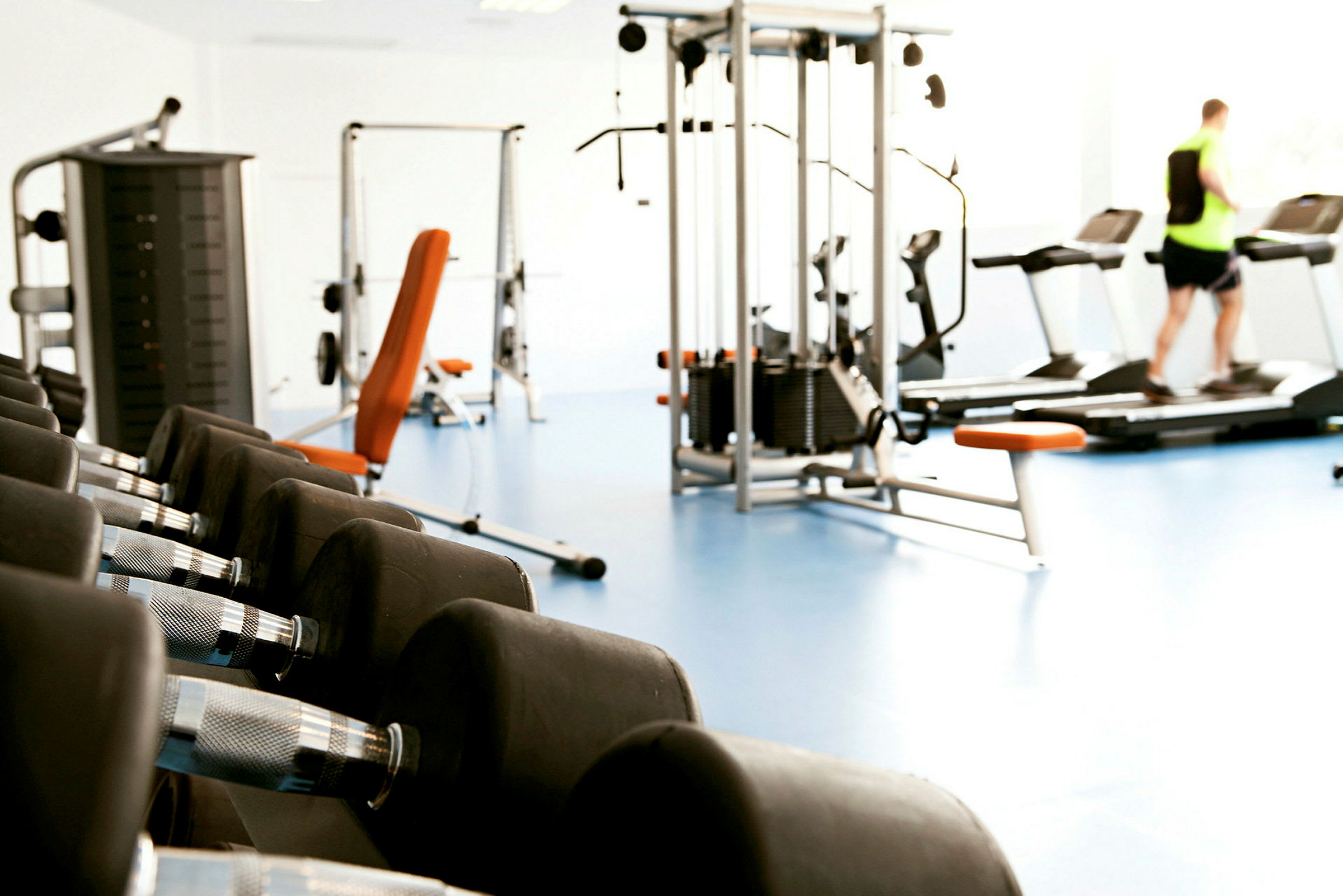 Et træningscenter med fitnessmaskiner, håndvægte og en person på et løbebånd. (AI-genereret tekst)