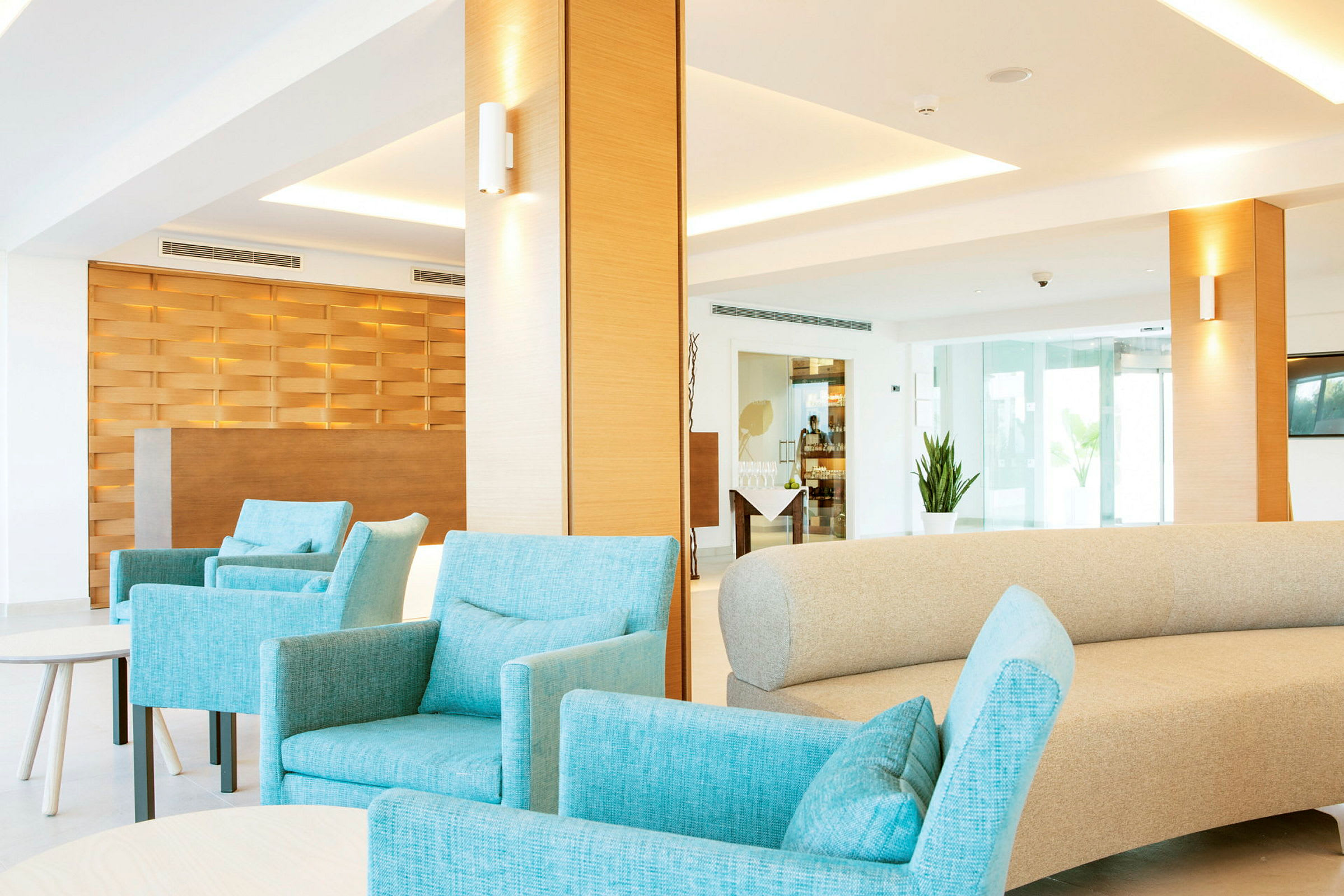 Interiør fra en moderne lounge med turkise lænestole og en beige sofa. Baggrunden viser en reception med en træbeklædning og store vinduer. (AI-genereret tekst)