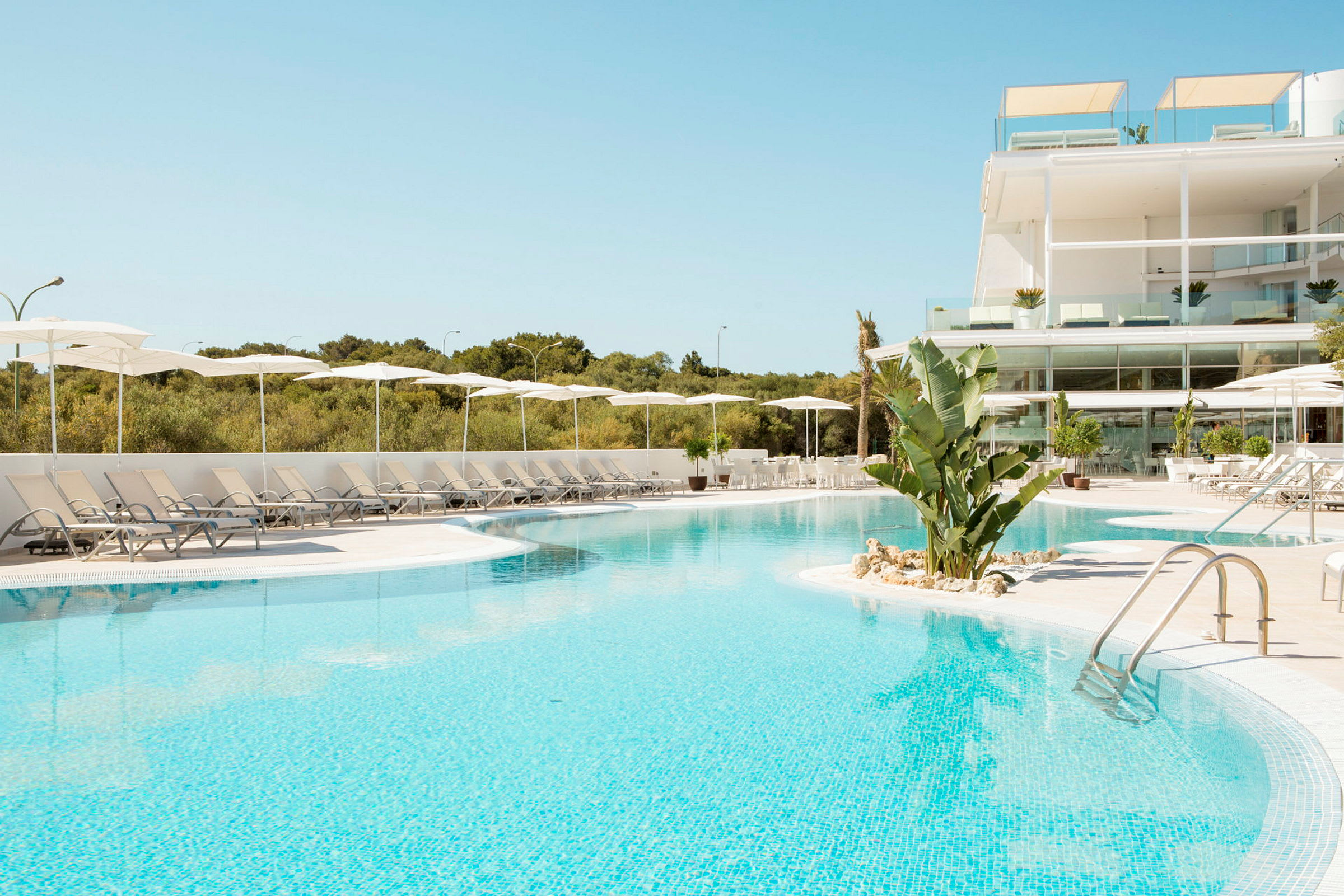 En stor udendørs pool omgivet af liggestole og parasoller. I baggrunden ses et moderne hotel. (AI-genereret tekst)