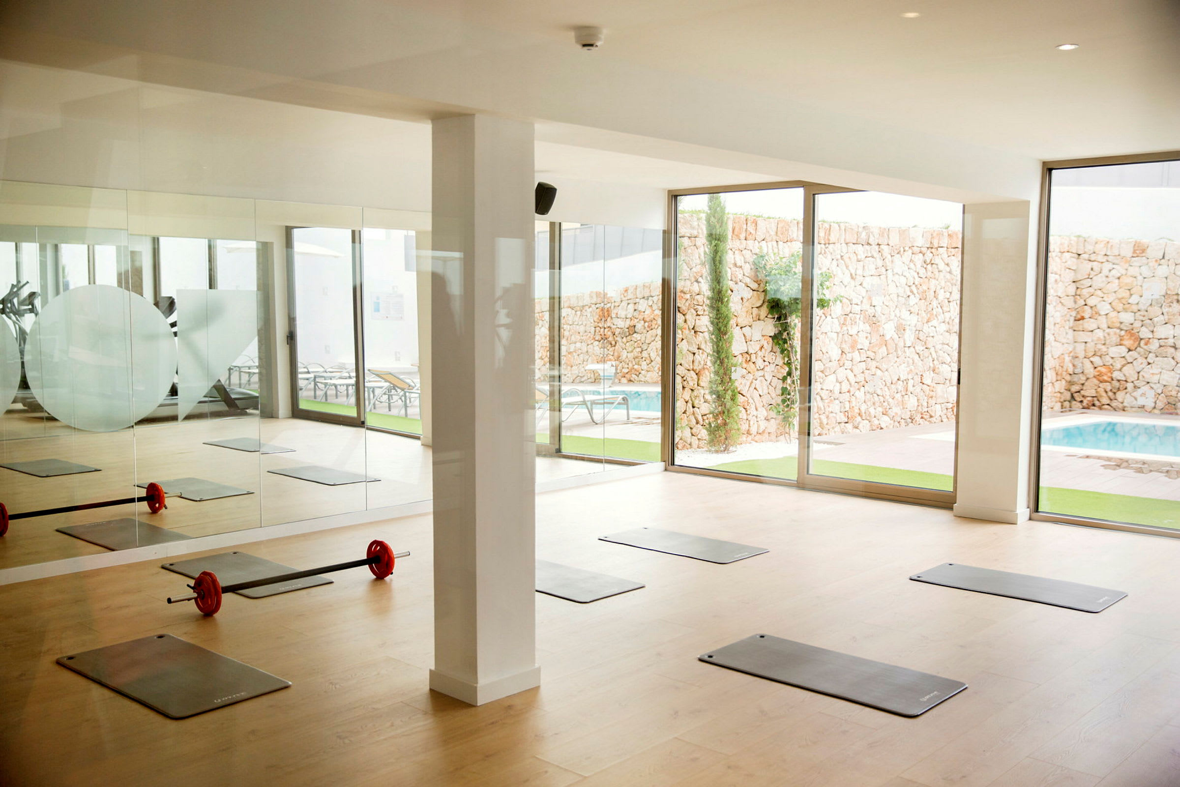 Et træningslokale med yogamåtter og en vægtstang på gulvet nær store glasvinduer mod en pool og en stenvæg. (AI-genereret tekst)