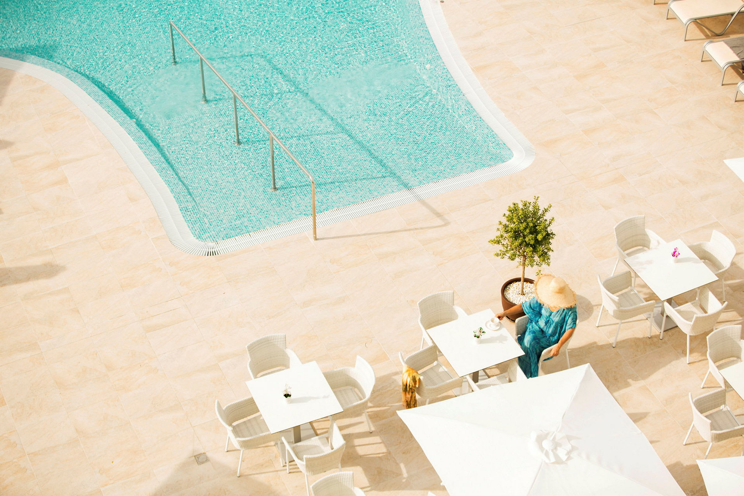 Udsigt over en pool med klart vand. På en solterrasse er der borde og stole, hvor en person sidder ved et bord med en kop. Personens ansigt dækkes af en solhat. (AI-genereret tekst)