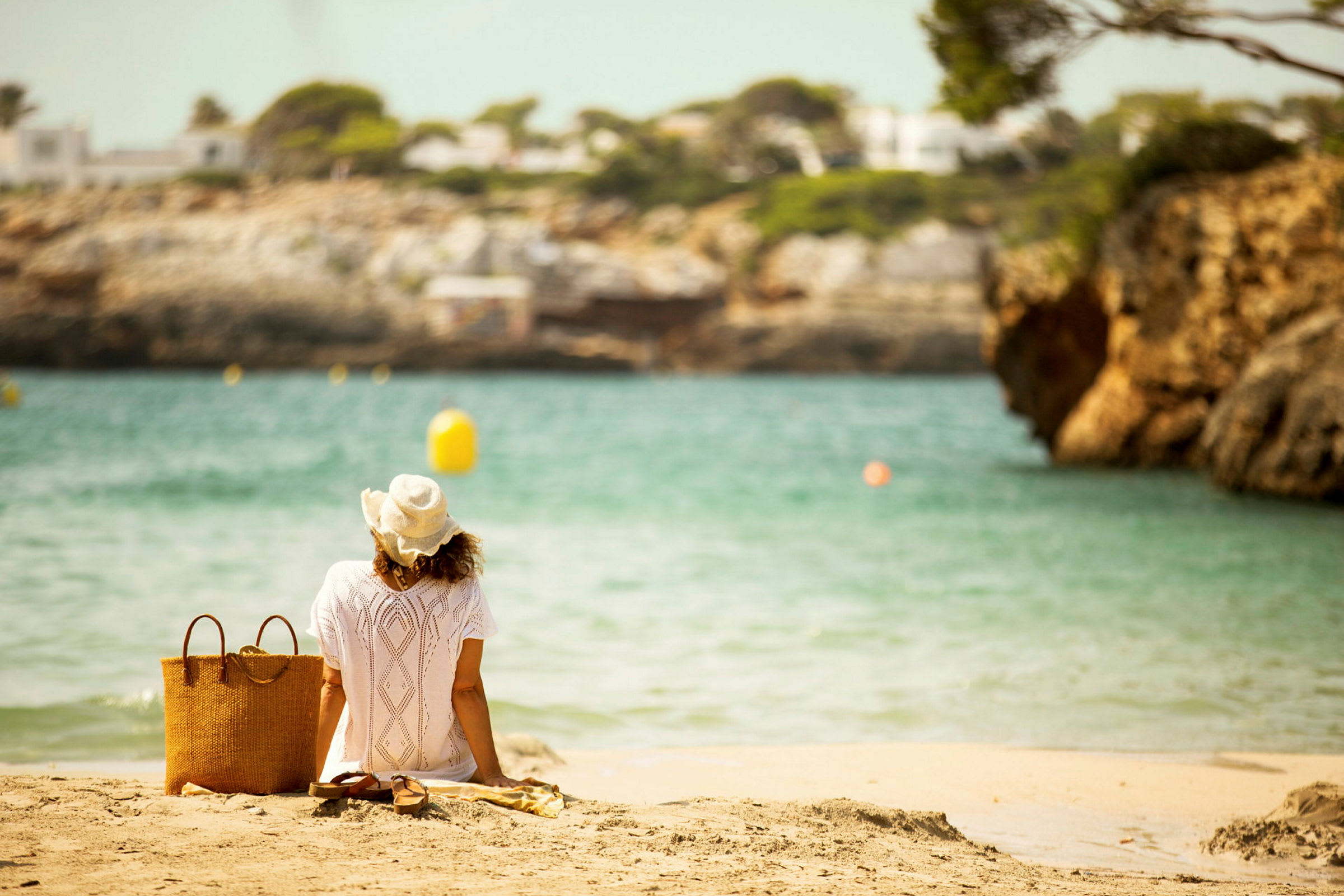 En person i en hvid kjole og hat sidder på en strand med udsigt over vandet. Ved siden af personen er der en kurv og et par sandaler. (AI-genereret tekst)