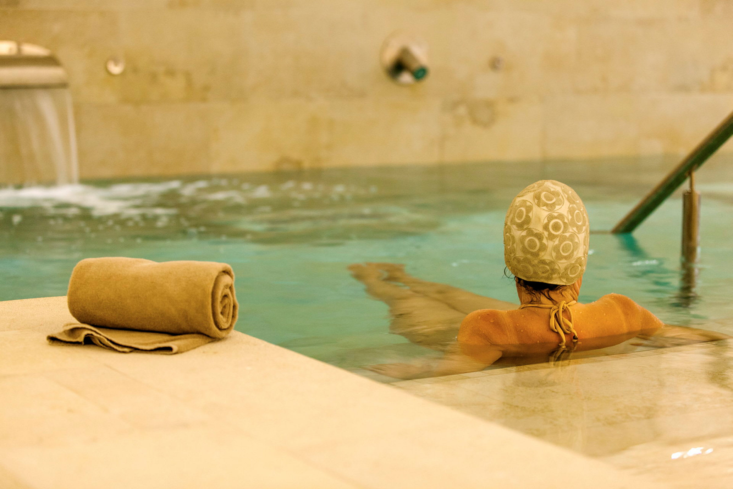 En person med badehætte sidder i en pool, med et sammenrullet håndklæde på kanten. (AI-genereret tekst)