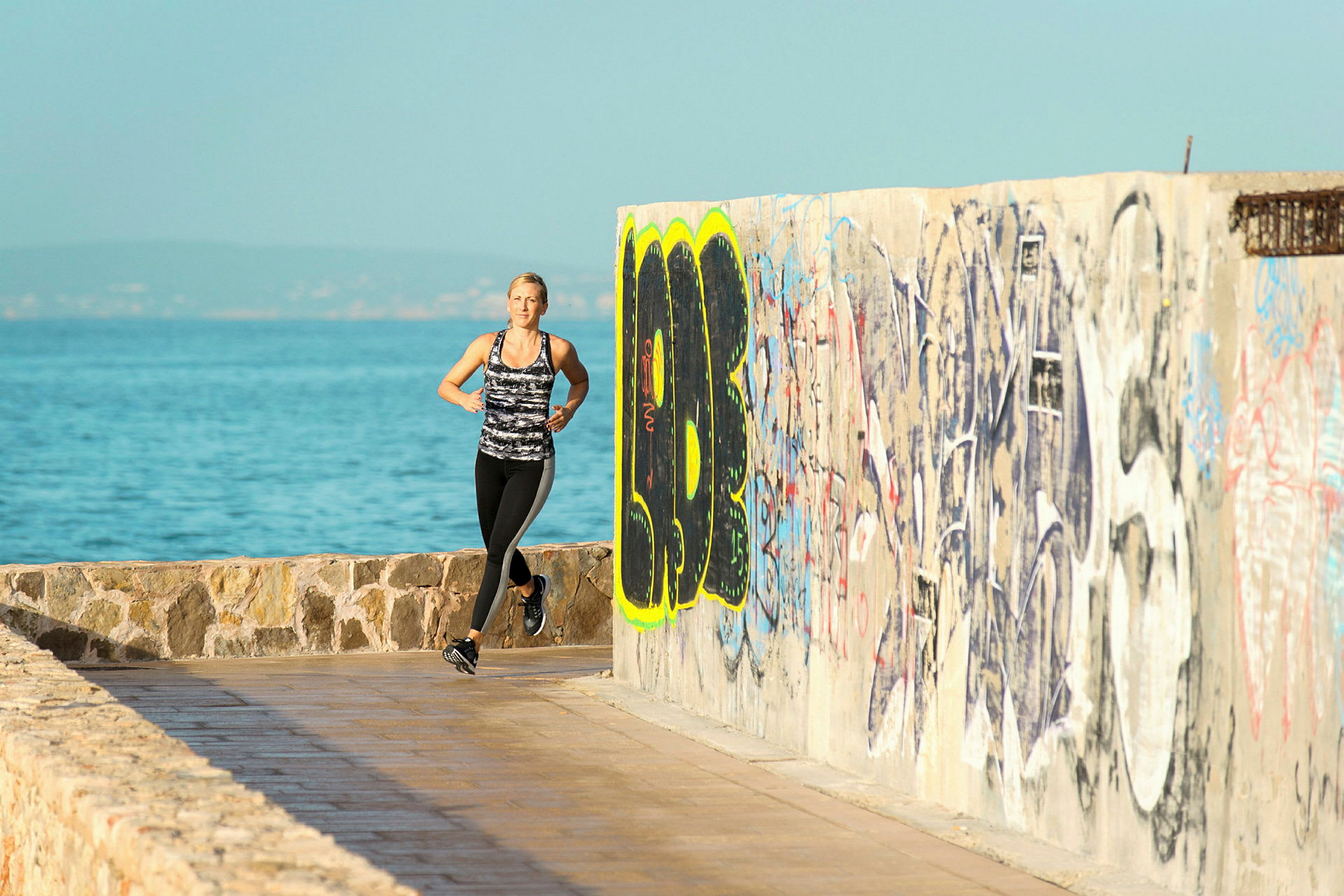 Person jogger langs en strandpromenade med havet i baggrunden og en væg malet med graffiti. (AI-genereret tekst)